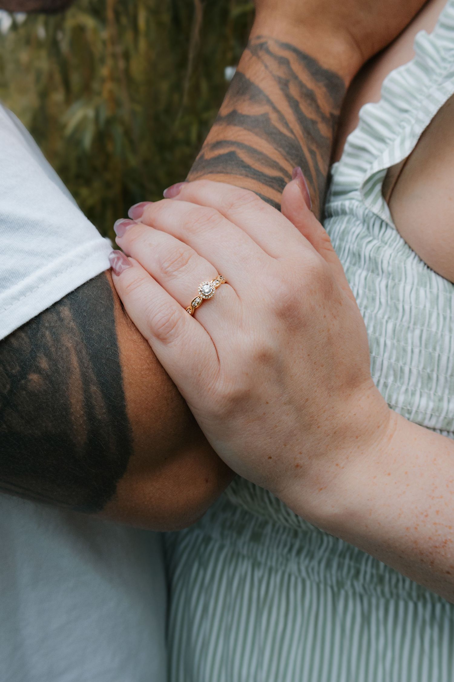 Close-up of a woman's hand wearing a gold engagement ring, resting on a tattooed man's arm.