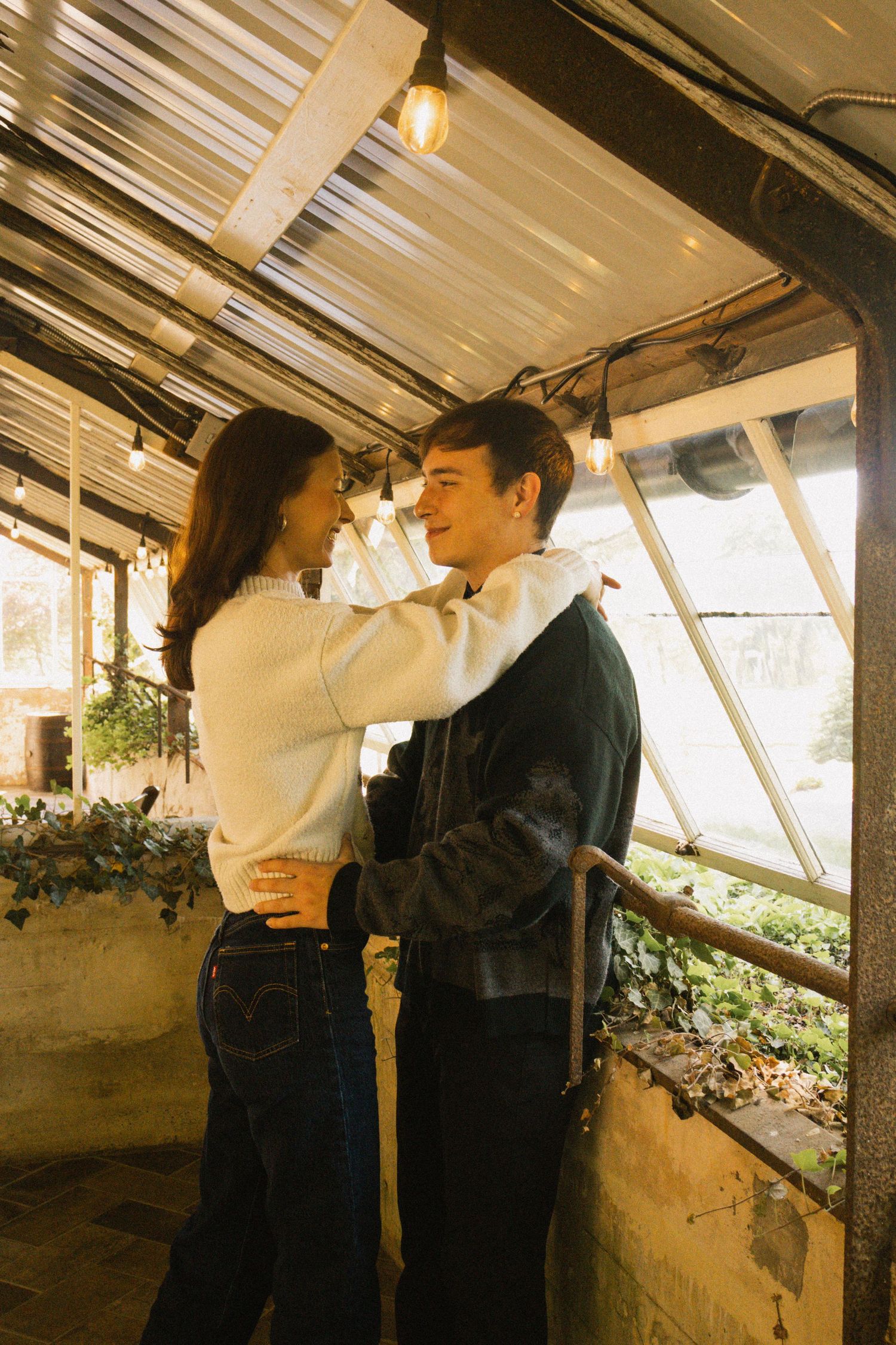 A couple embracing and smiling at each other inside a warmly lit greenhouse with string lights and green plants.