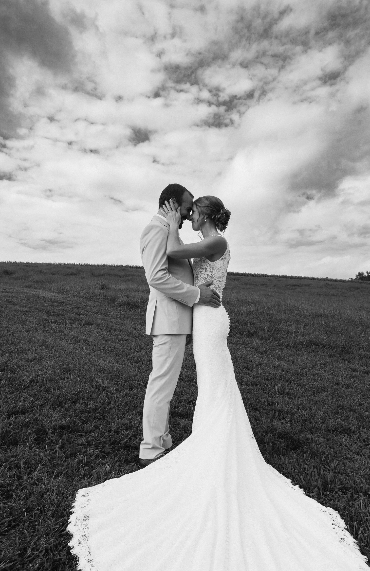 Black and white photo of a bride and groom embracing in a field under a cloudy sky, wedding dress train spread on grass.