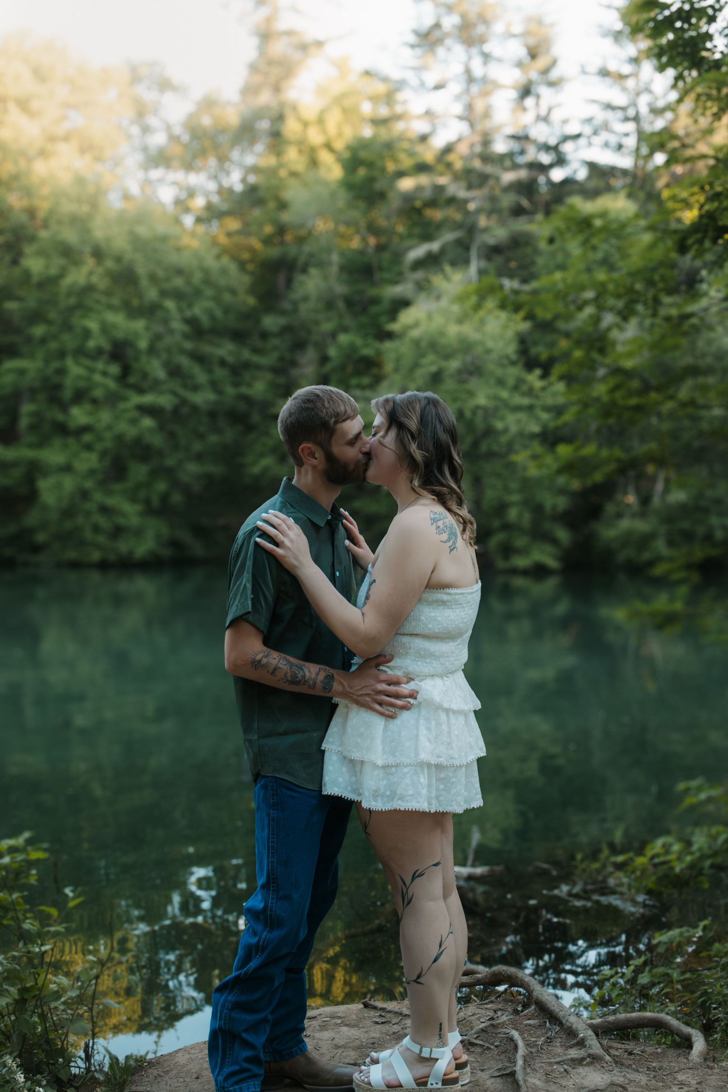 Couple sharing a romantic kiss by a serene green lake surrounded by lush forest trees during golden hour.