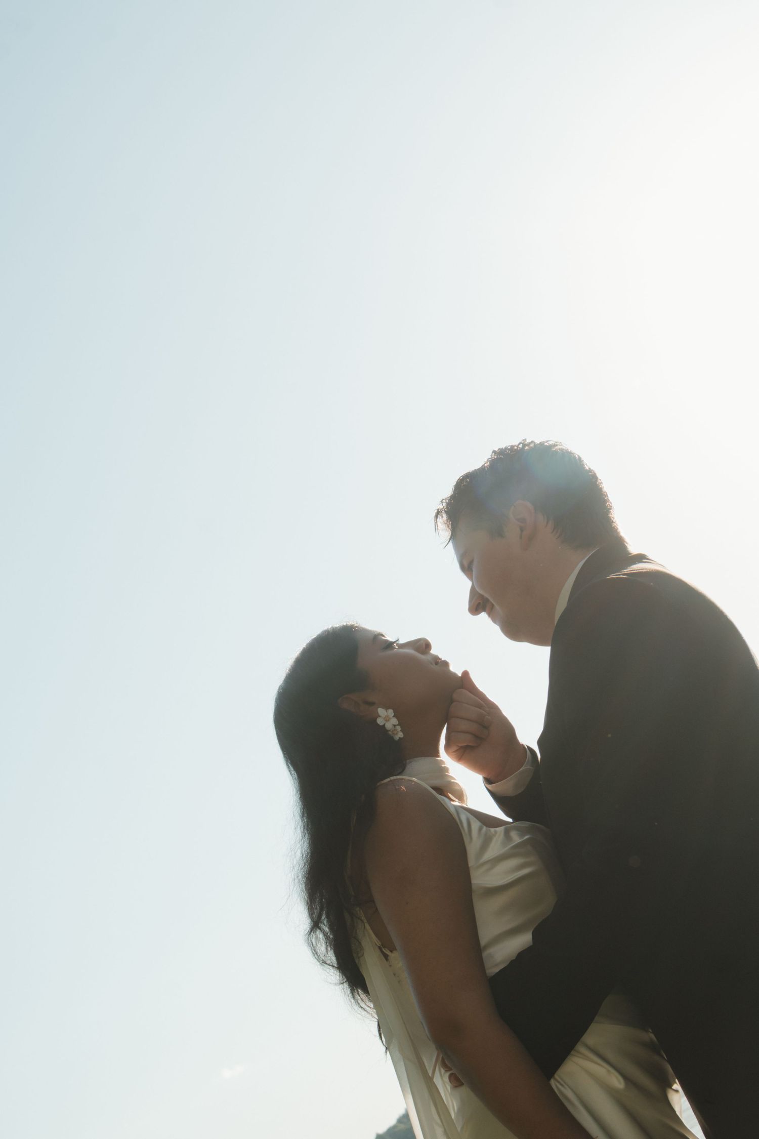 A couple shares a romantic moment outdoors, backlit by bright sunlight against a clear sky, in wedding attire.
