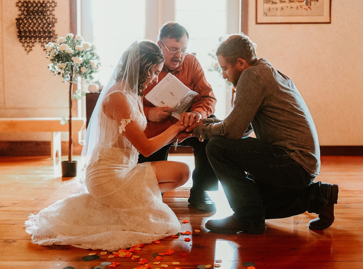 Couple in wedding attire sits on floor during intimate indoor ceremony moment.