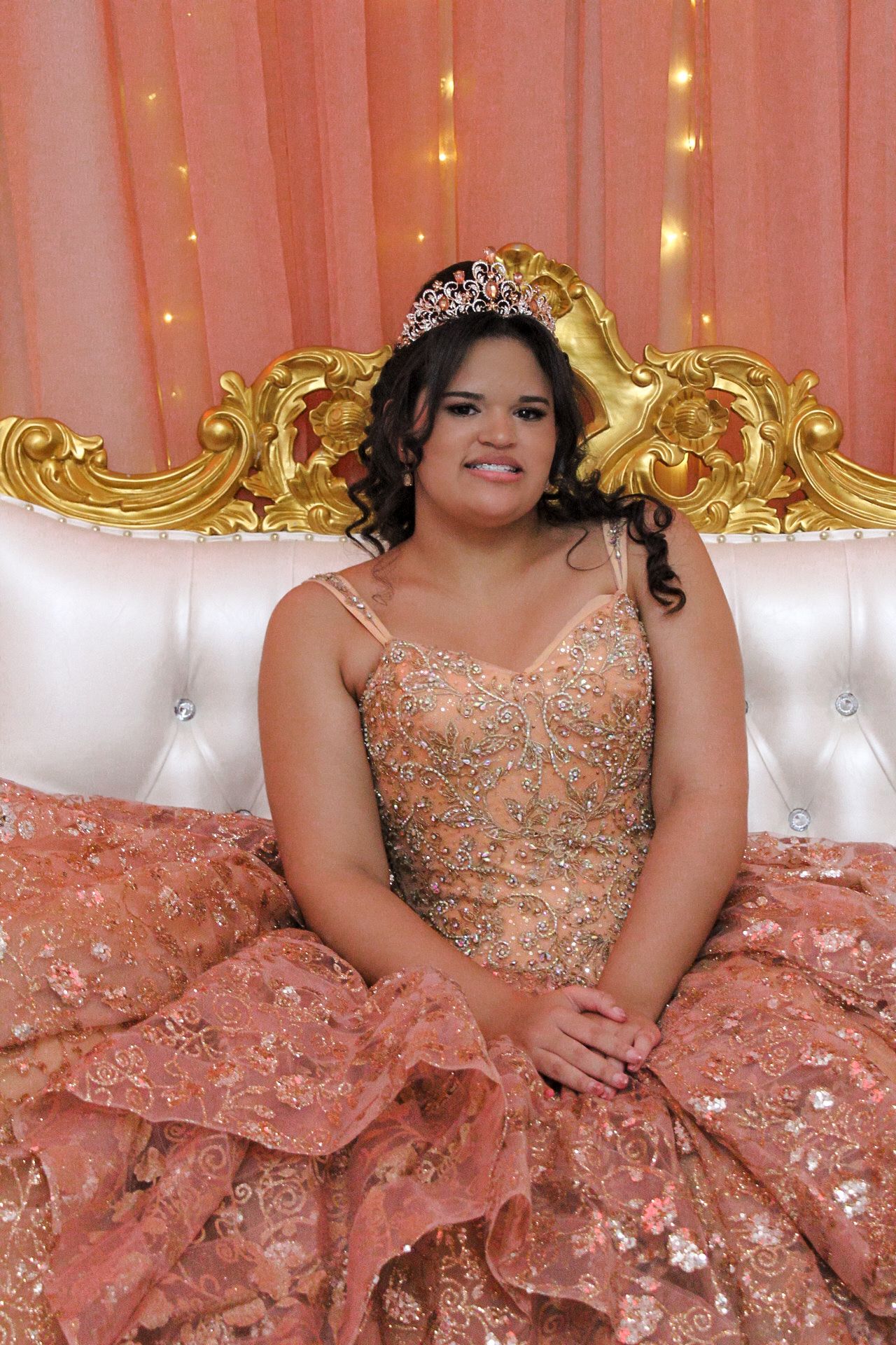 Portrait of guest in beaded rose gold quinceañera gown posing on ornate white and gold throne.