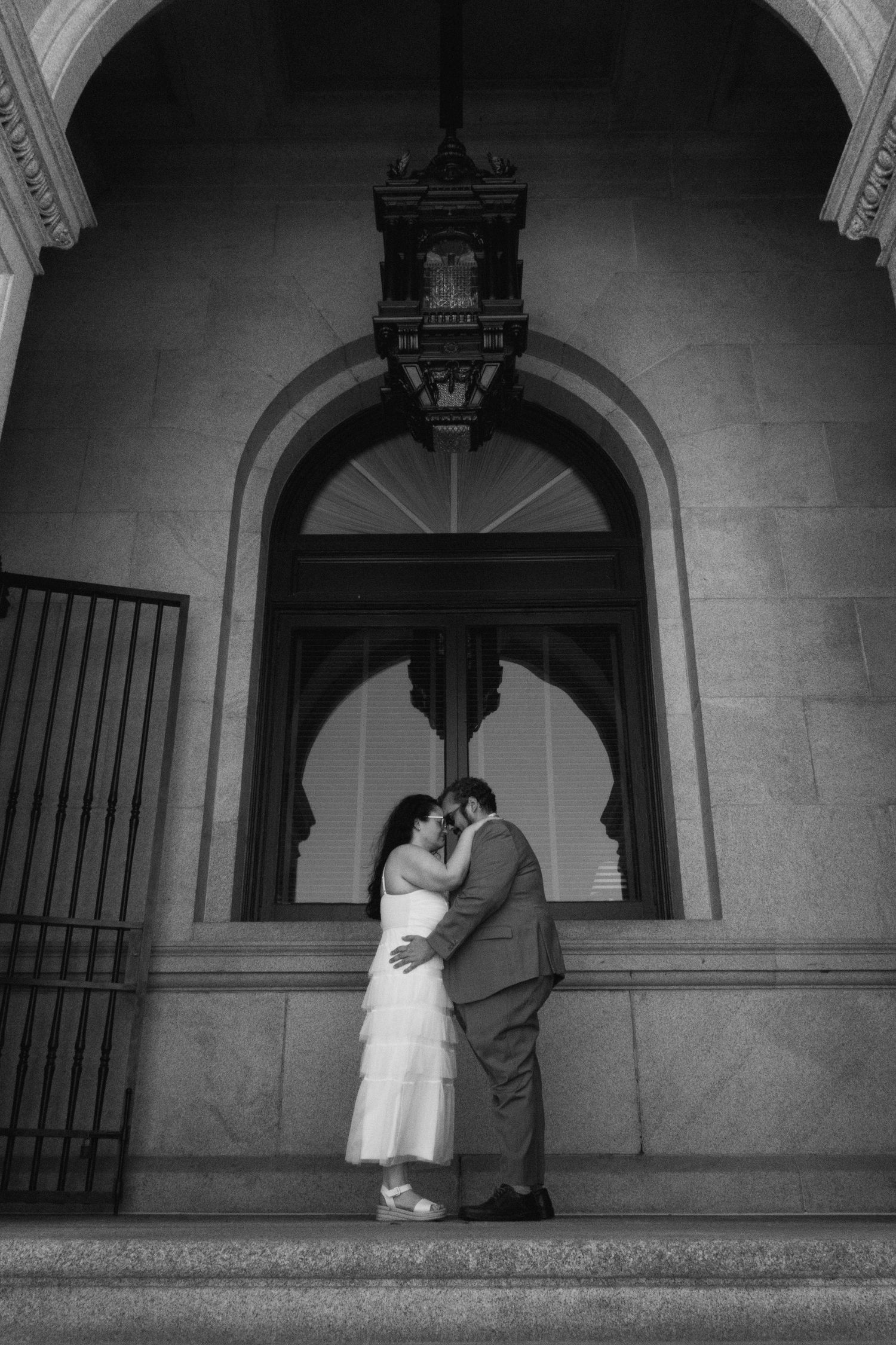 Black and white photo of a couple sharing a romantic kiss on steps beneath an ornate stone archway.