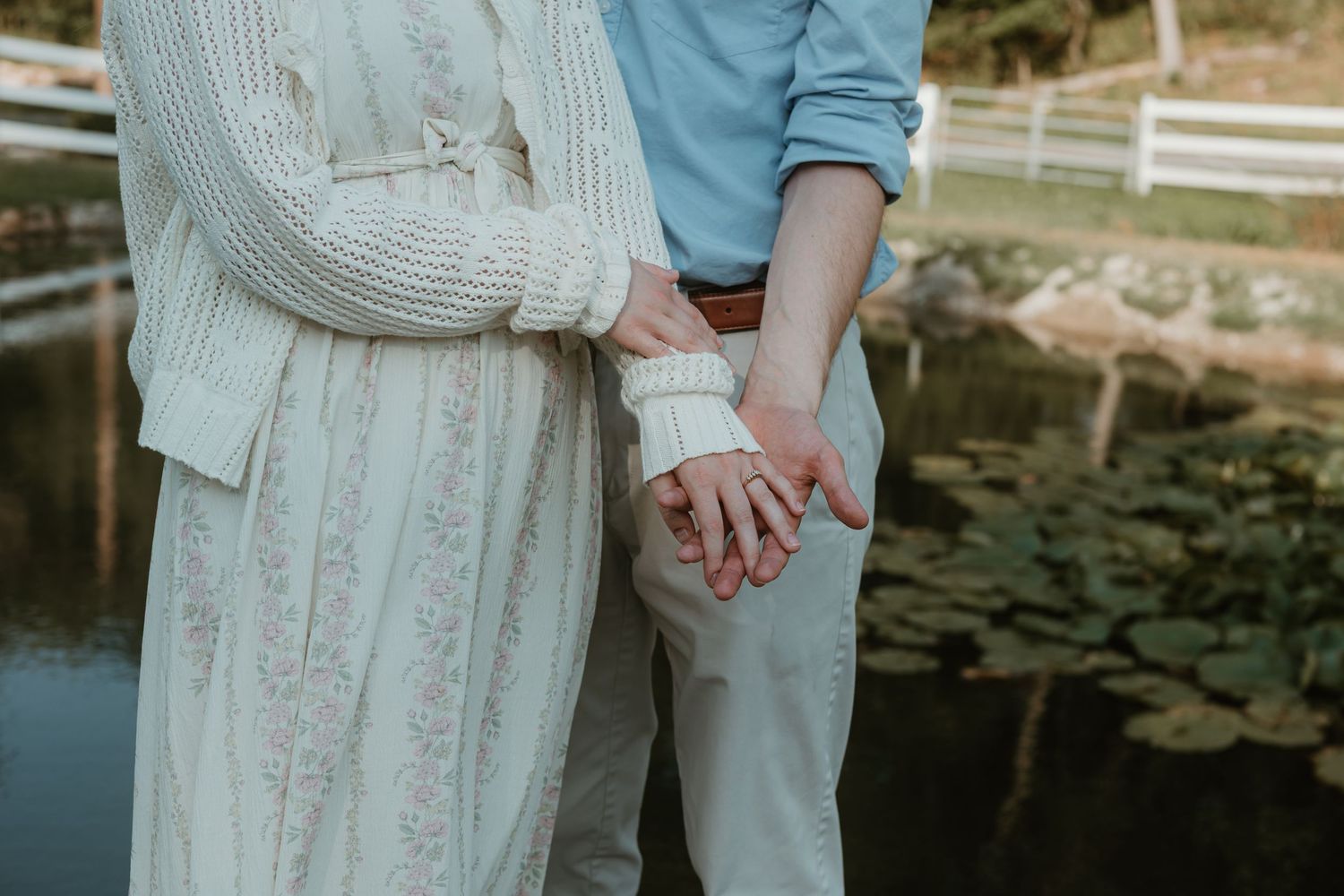 Couple holding hands near a serene pond, woman in white lace dress and man in blue shirt and khaki pants.