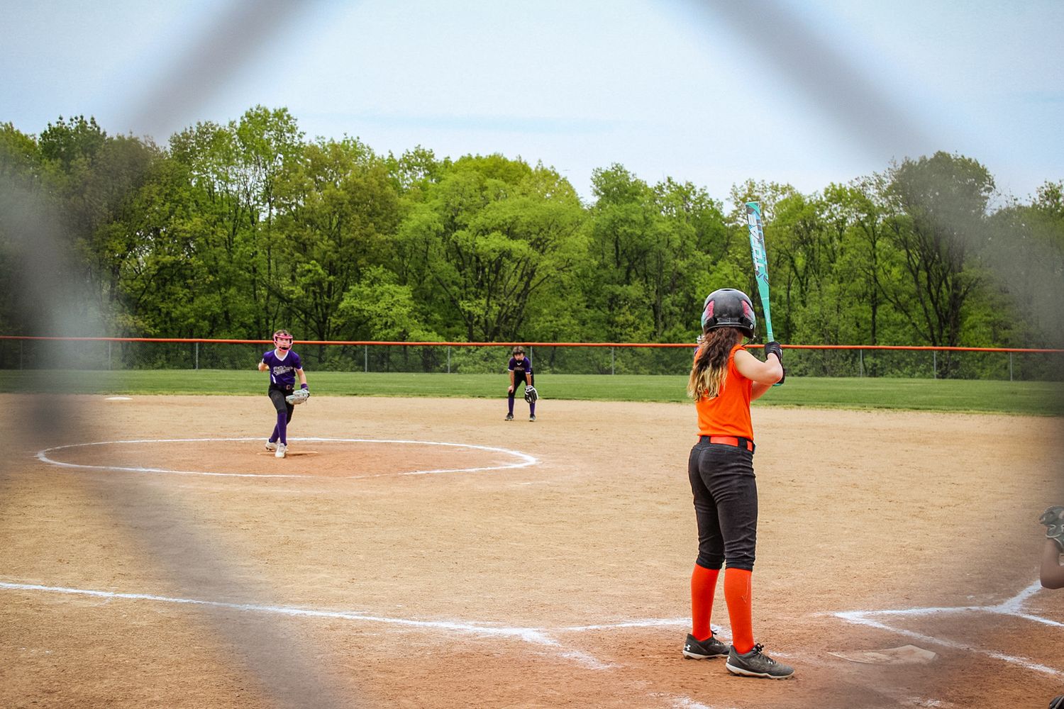Softball player in orange uniform at bat on dusty field during game.