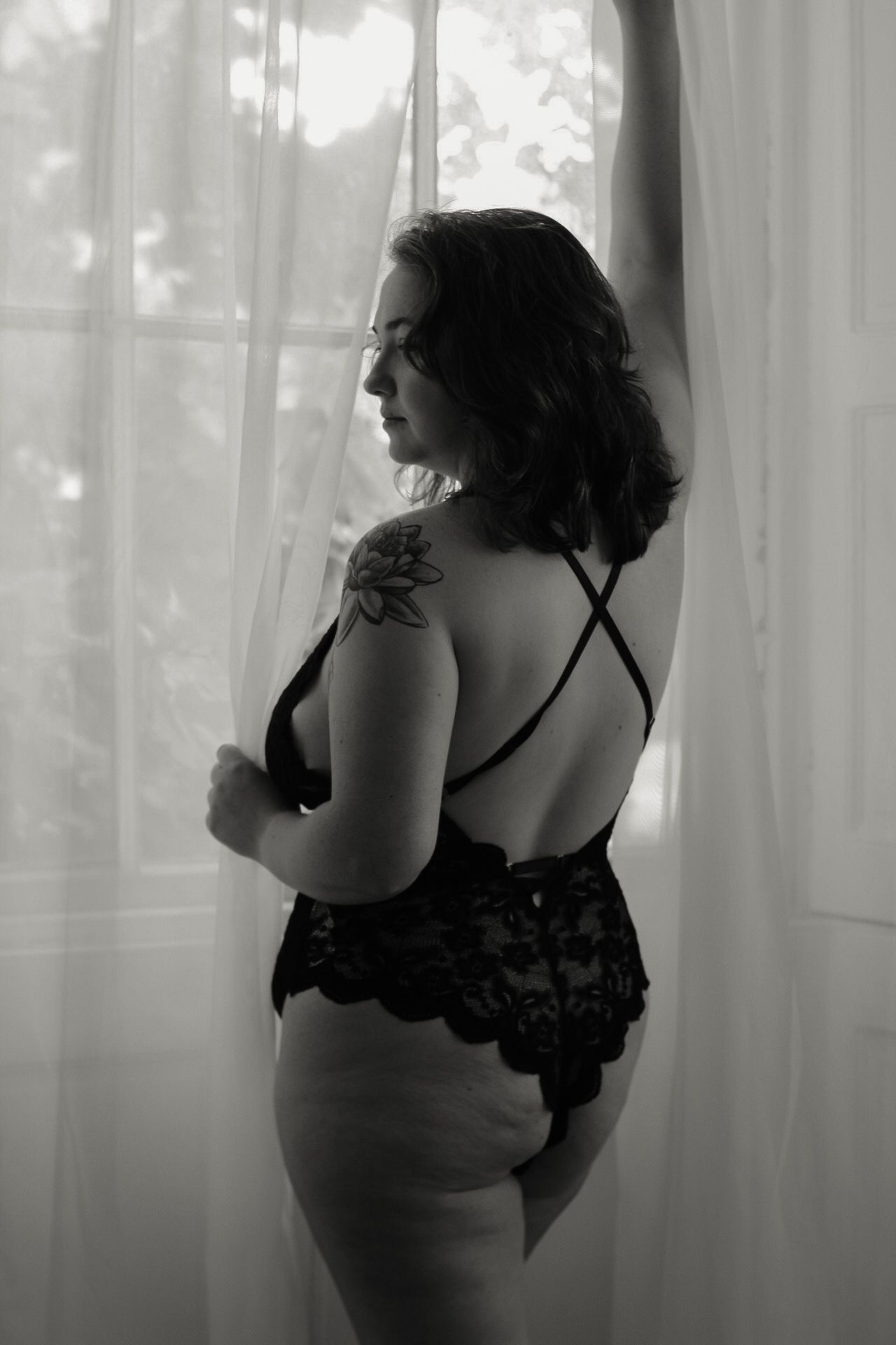Black and white portrait of model in lace lingerie showing shoulder tattoo by window.
