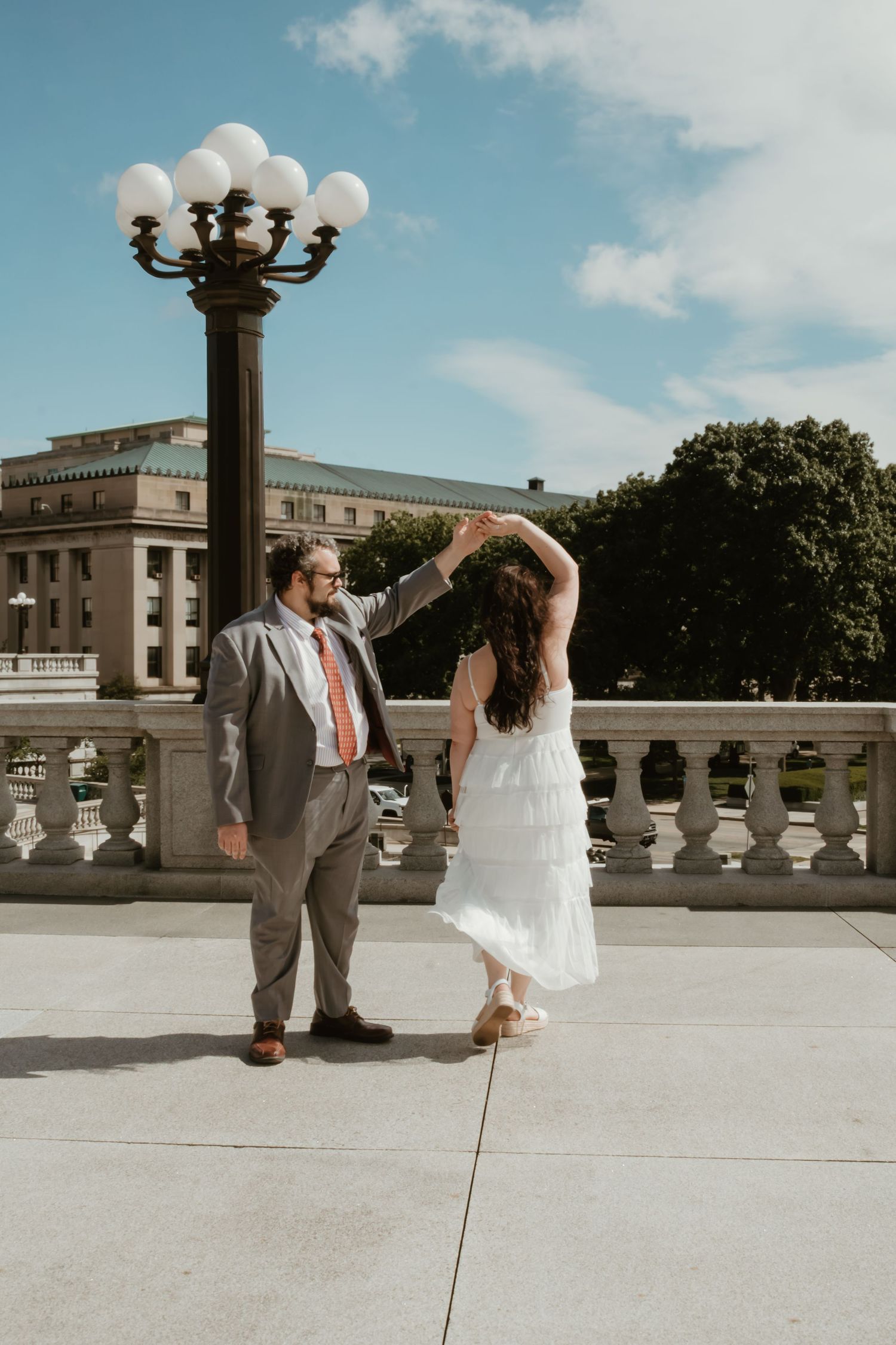 A couple dances outdoors on a terrace, the groom in a gray suit and the bride in a white dress, with a lamp post behind them.