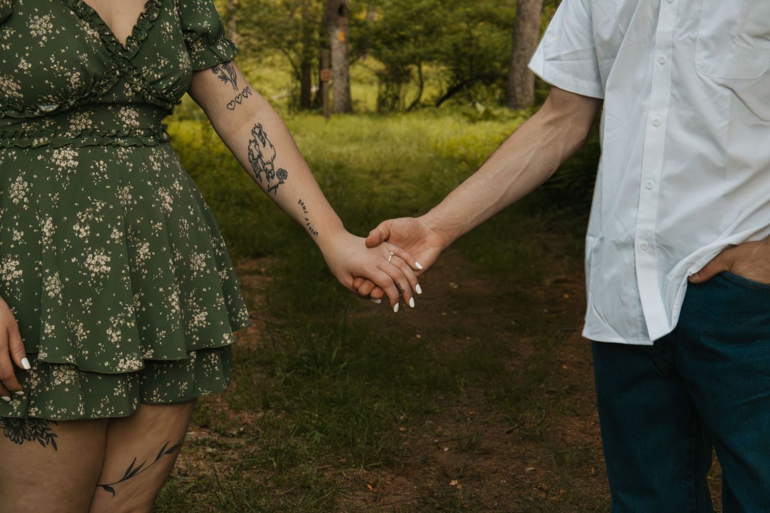 Couple holding hands outdoors in a park, woman wearing green floral dress with tattoos on her arm and leg.