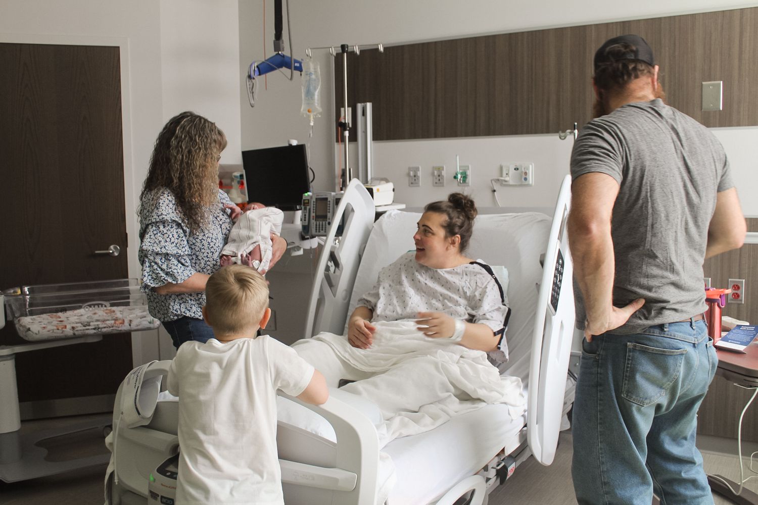 Family members gathered in a bright hospital room around a bed.