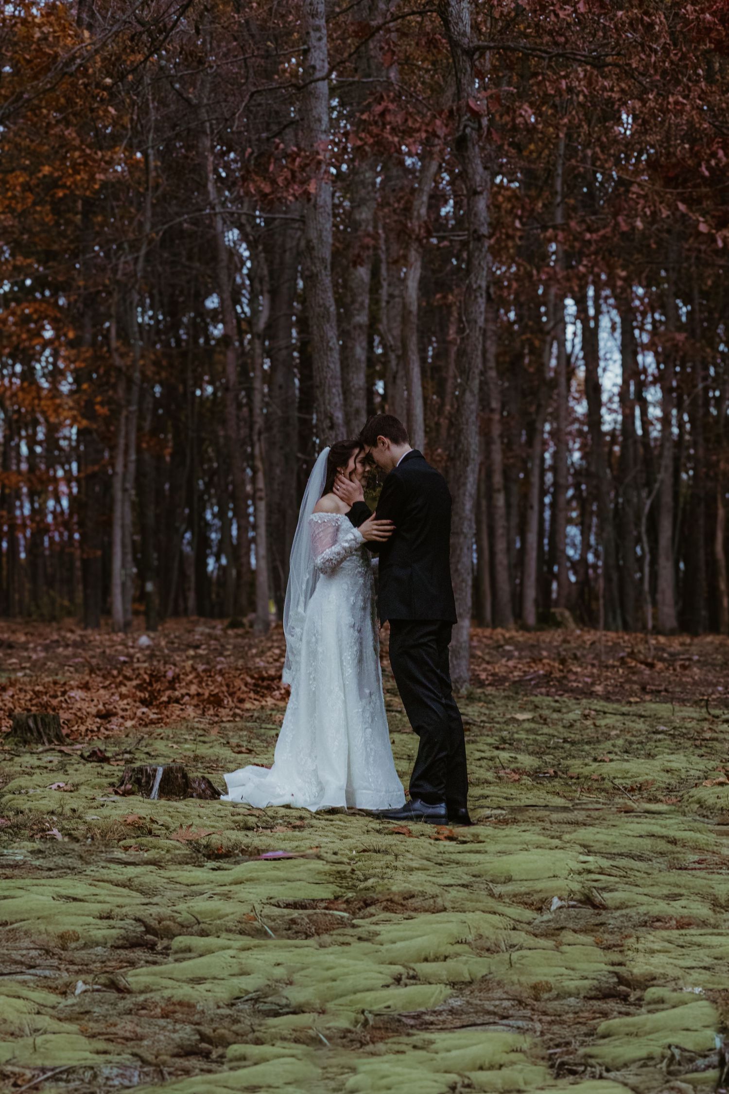 Newlyweds embrace on mossy ground surrounded by autumn trees in a moody forest setting.