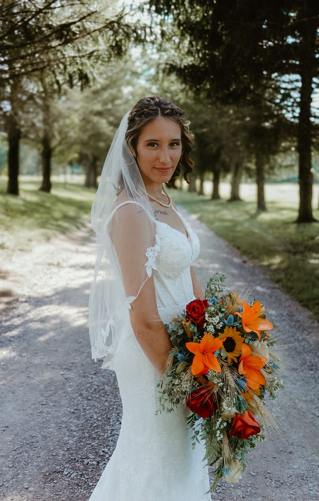 A bride in a white wedding dress holds a vibrant bouquet of sunflowers and red roses on a wooded path.