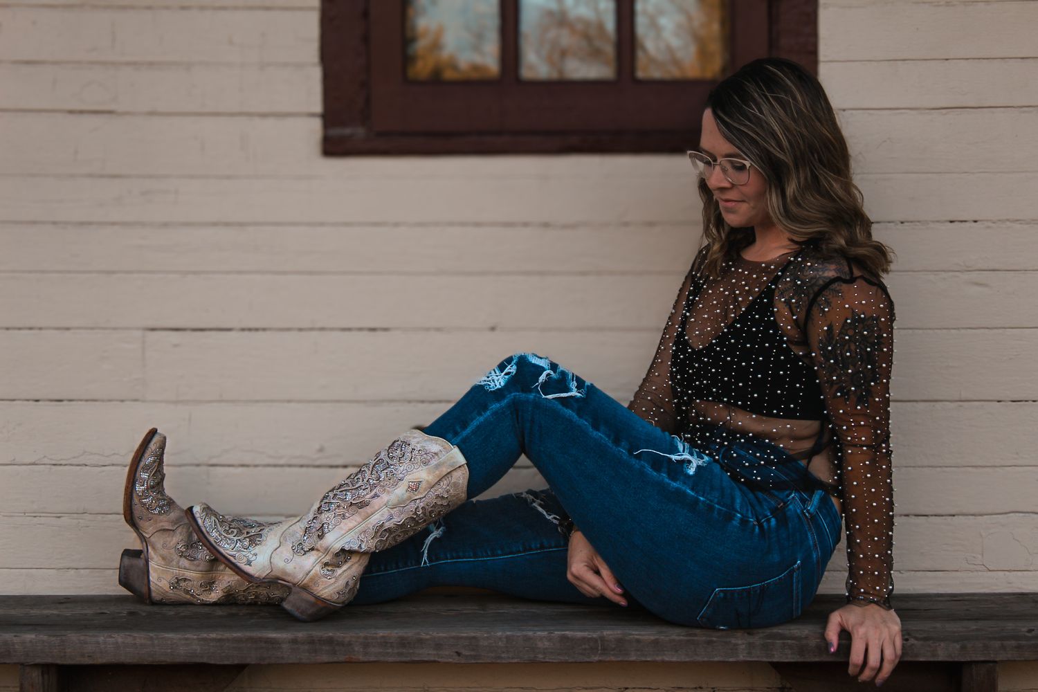 Relaxed pose on a wooden bench wearing western boots, ripped jeans, and a sheer mesh top.