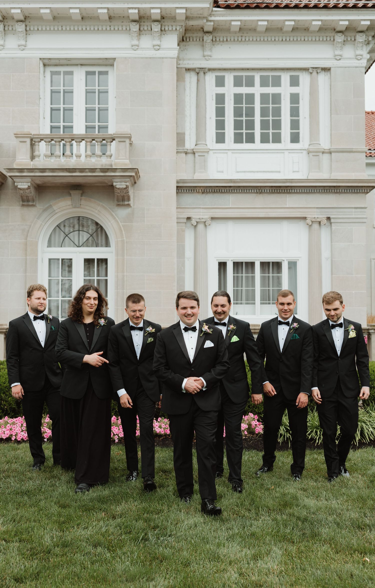 Groom and groomsmen in black tuxedos walking together in front of an elegant stone mansion with pink flowers.