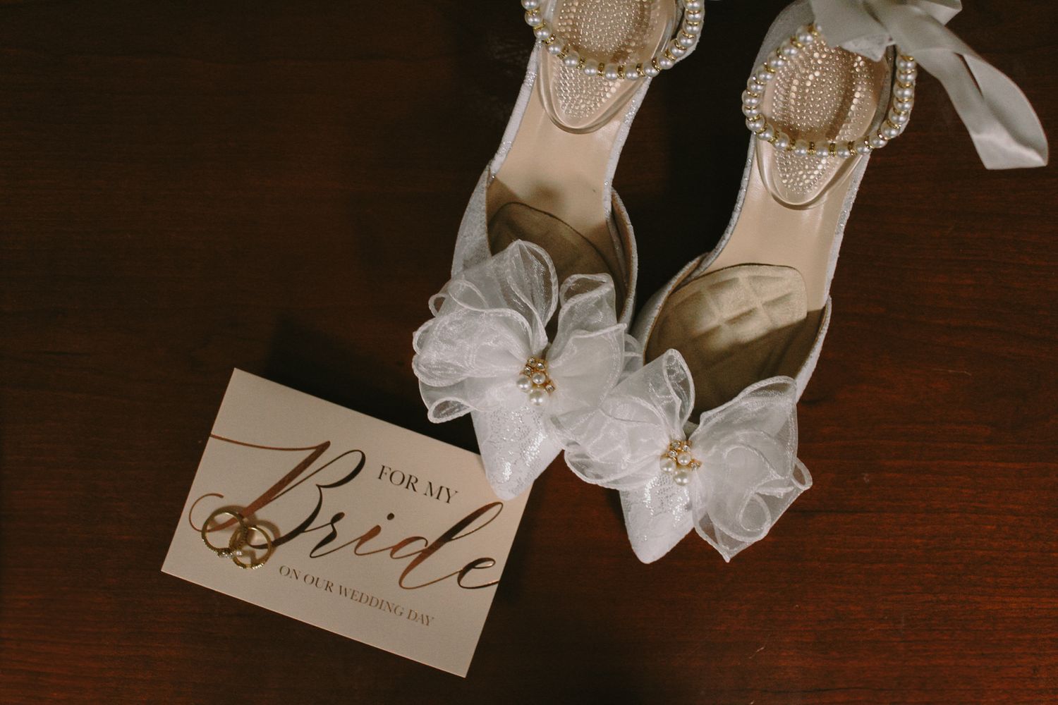 Elegant bridal shoes with white organza bows beside 'For my Bride' card on dark wooden surface.