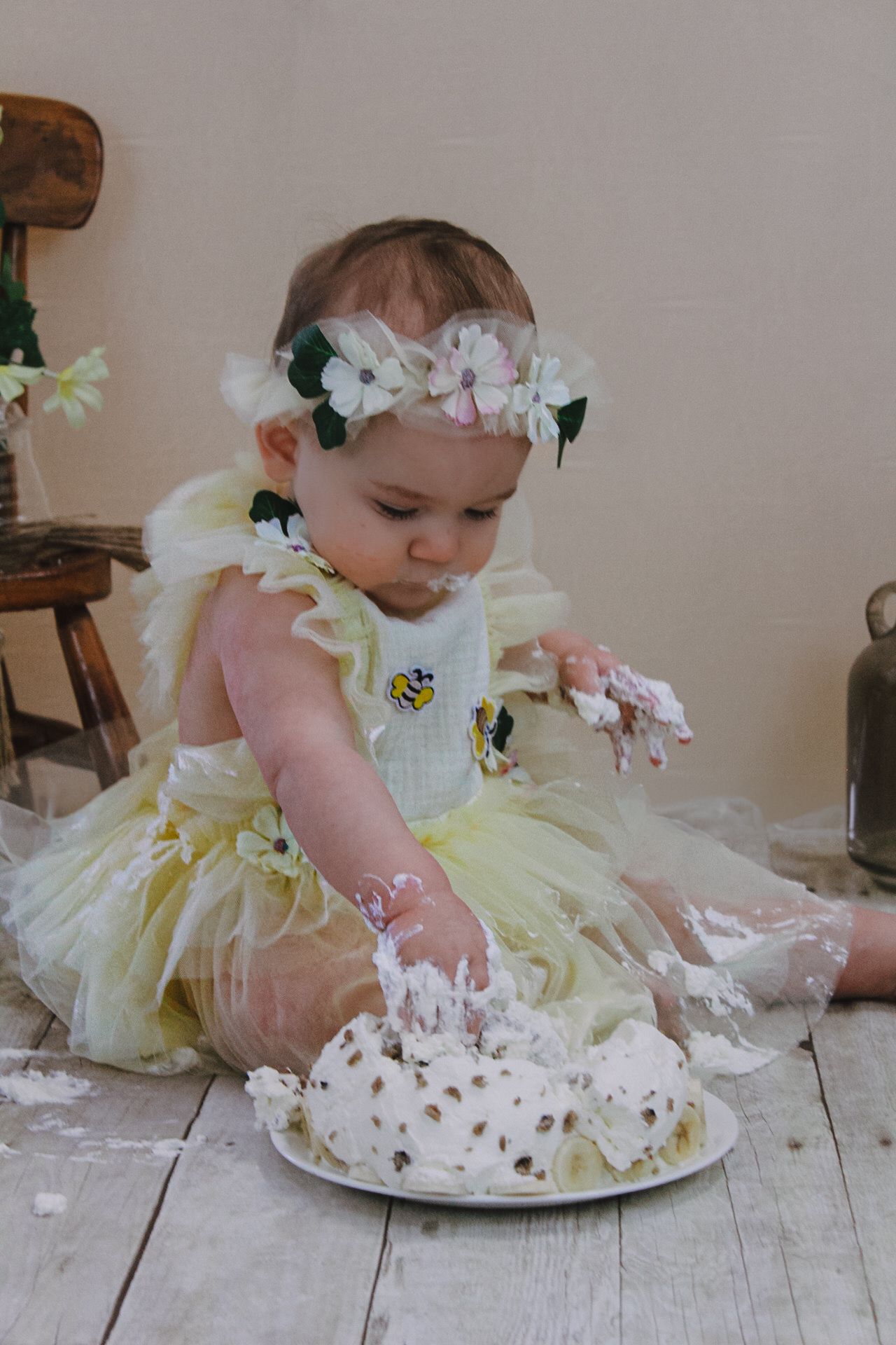 A baby in floral crown and yellow dress plays with frosting from a cake.