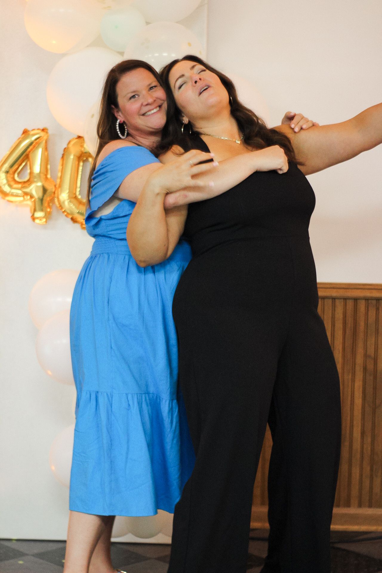 Friends laughing and embracing at a celebration with gold balloons visible.