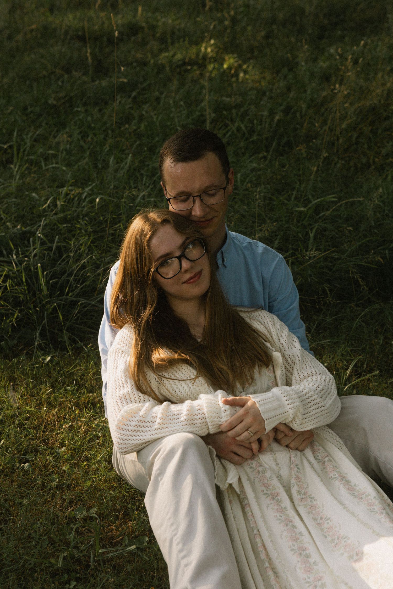 Couple sitting in green grass field, woman in white dress leaning back on man wearing glasses.