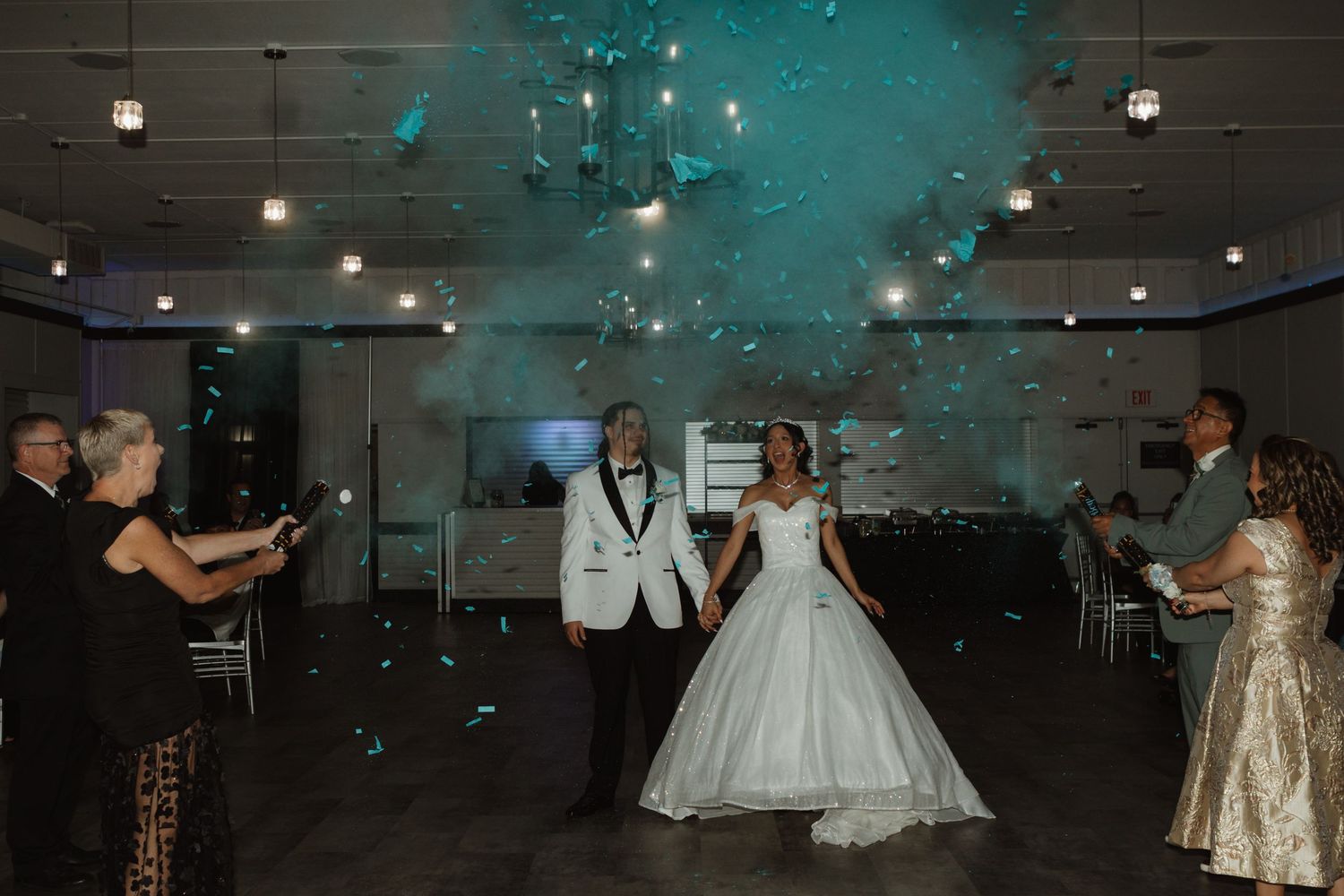 Bride and groom enter wedding reception hall with teal confetti falling from ceiling, guests celebrating around them.
