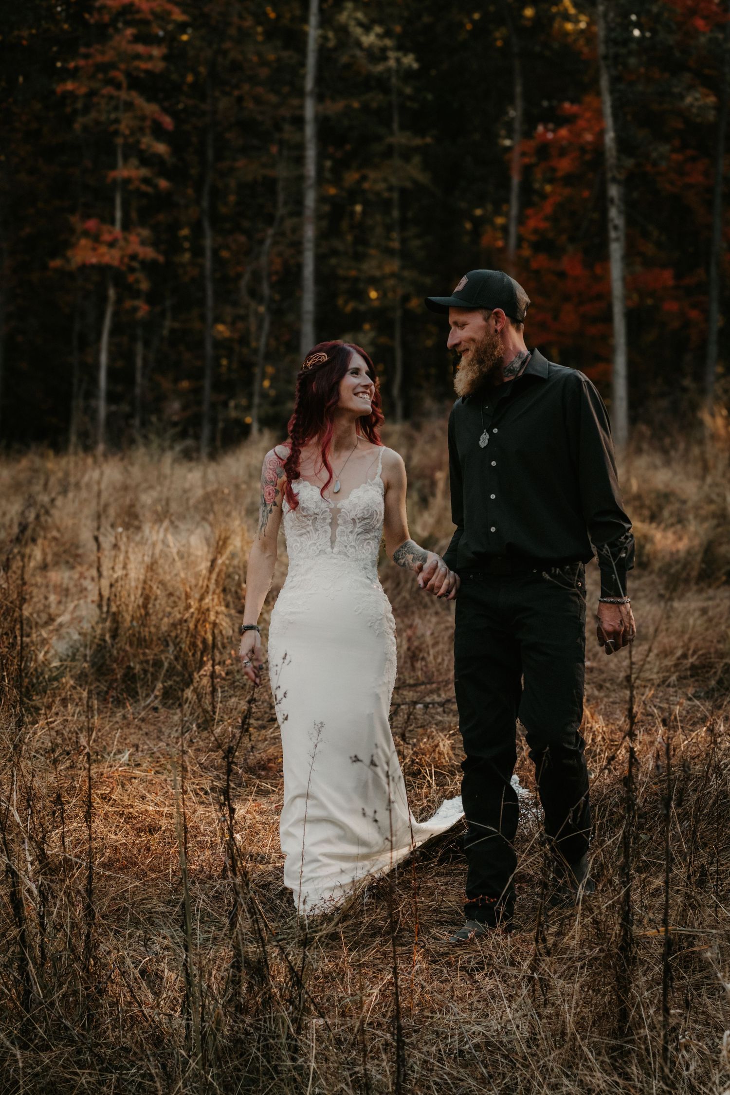 A couple in wedding attire walks through a golden autumn field with dark forest in the background during sunset.