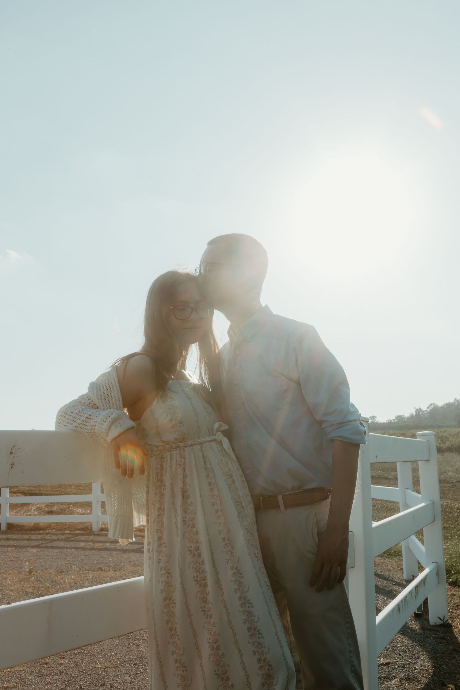 Couple sharing a tender moment by a white fence with bright sunlight creating a dreamy backlit effect.