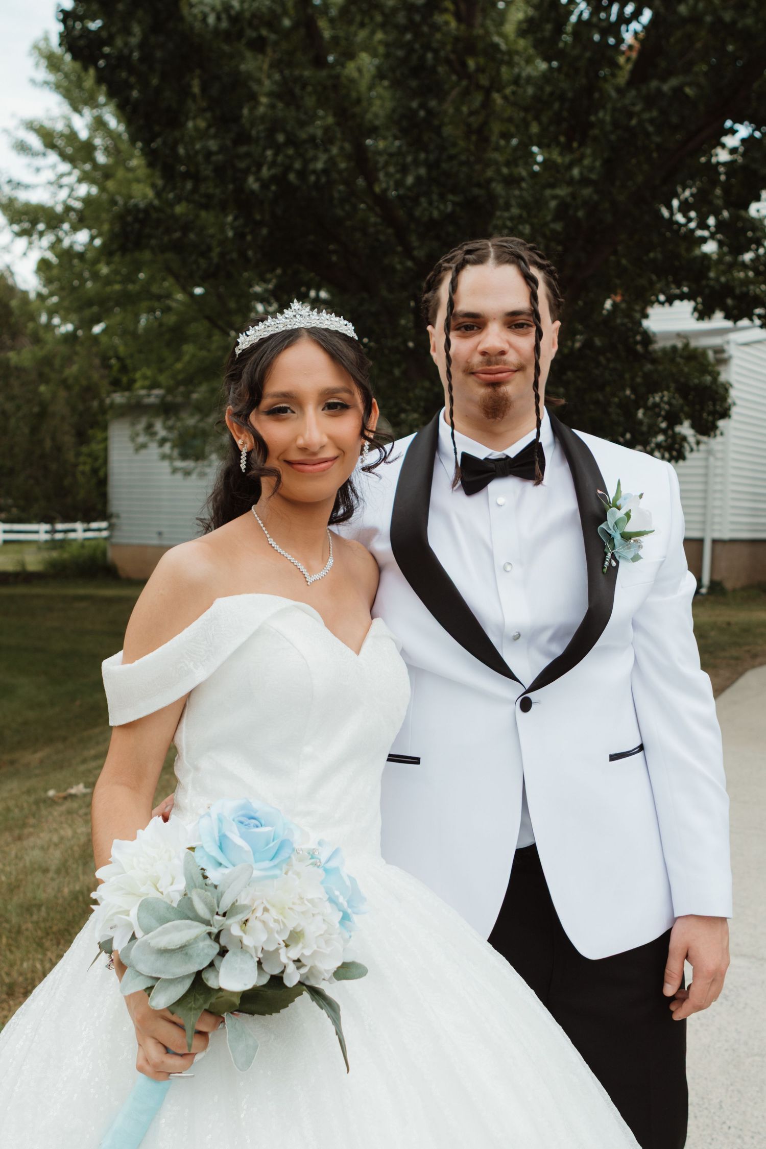 Bride in white ball gown and groom in white tuxedo posing together outdoors holding blue floral bouquet.