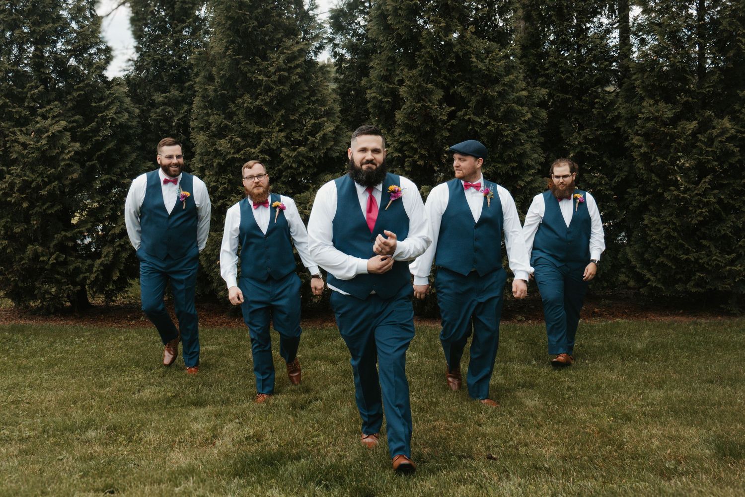 Five groomsmen in navy blue vests and pink ties walking confidently across green lawn at outdoor wedding.