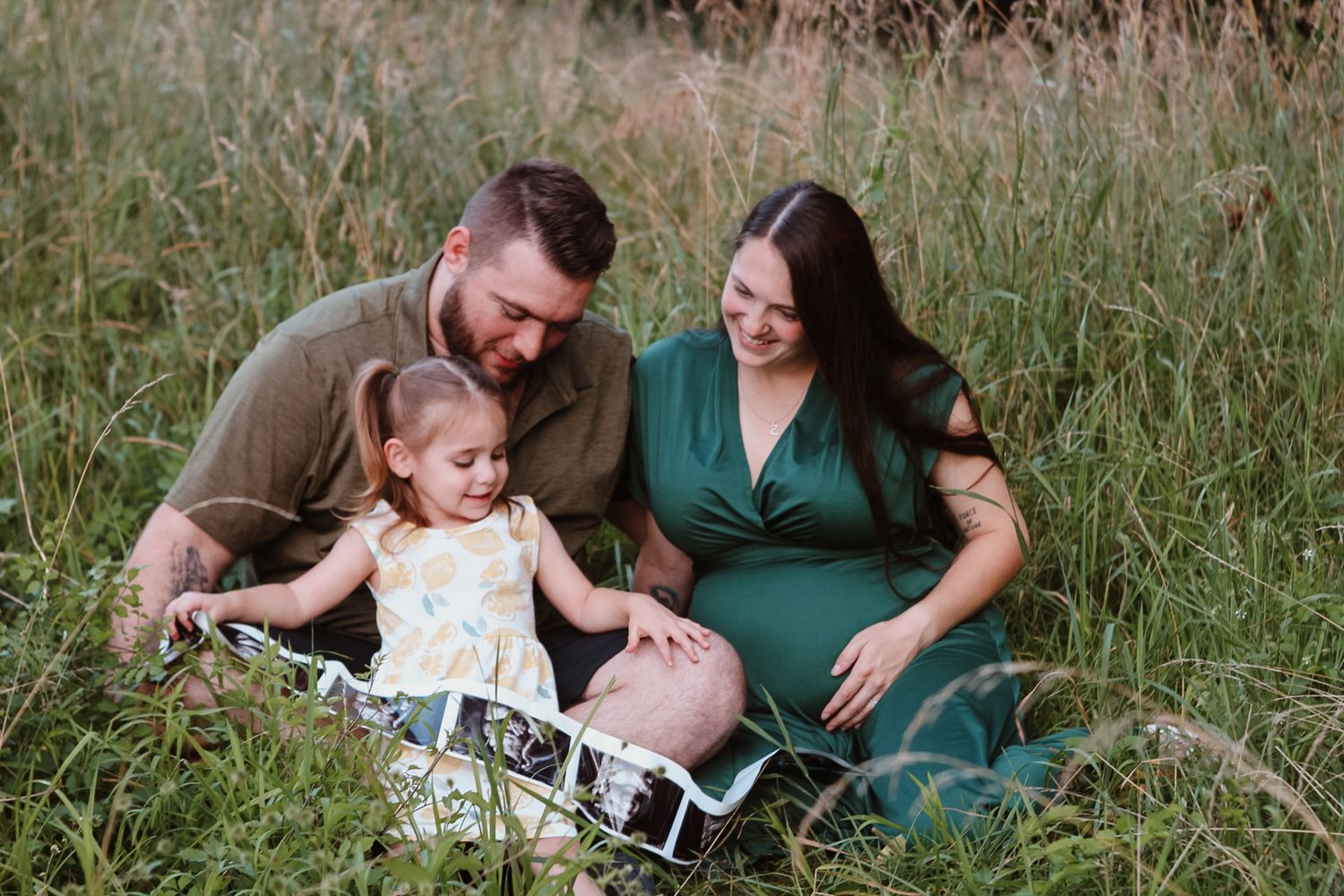 A family sits together in tall grass looking at ultrasound photos during a maternity photoshoot.