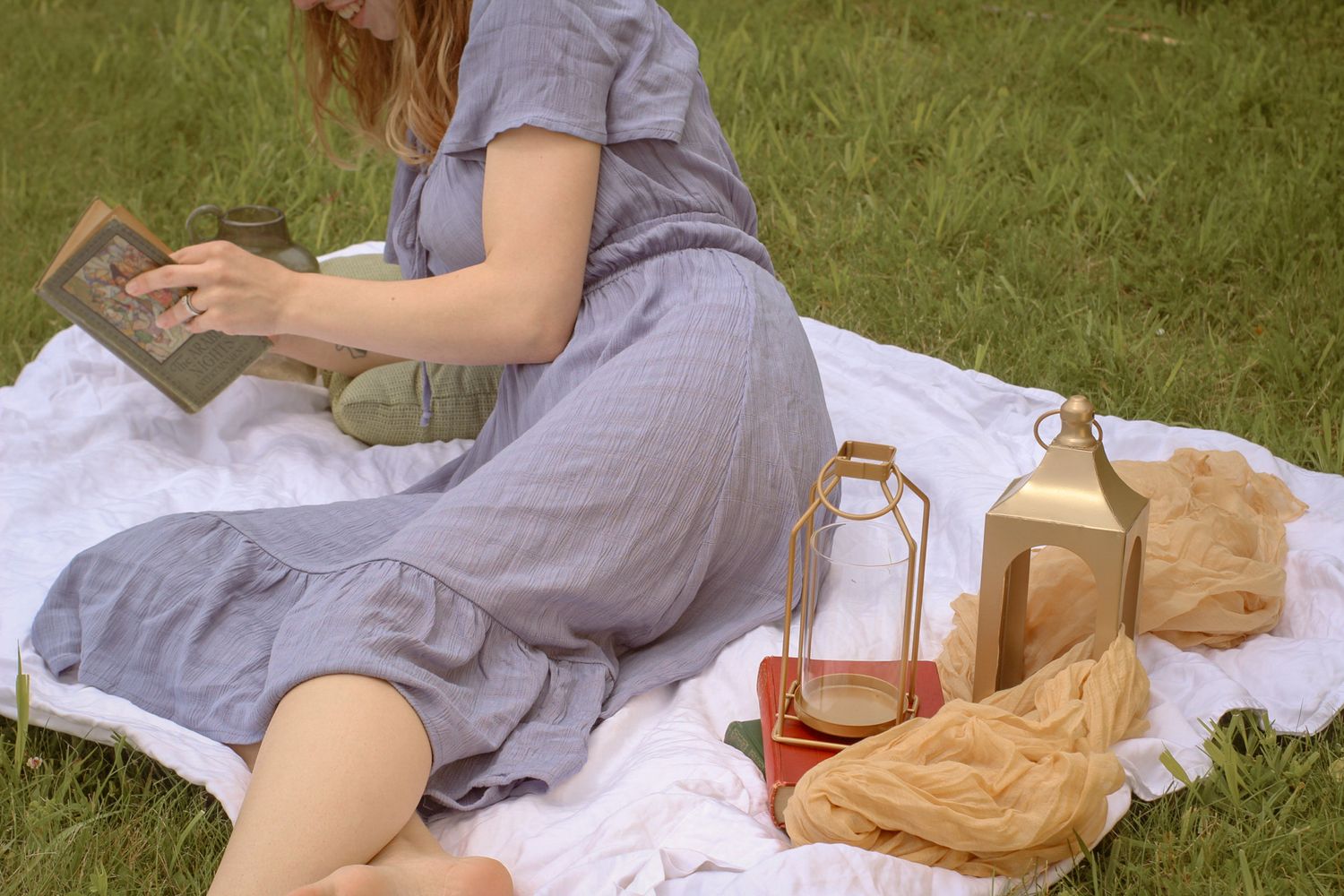Woman in a blue dress enjoys a picnic on a white blanket with lanterns and old vintage books. Cottage core vibes