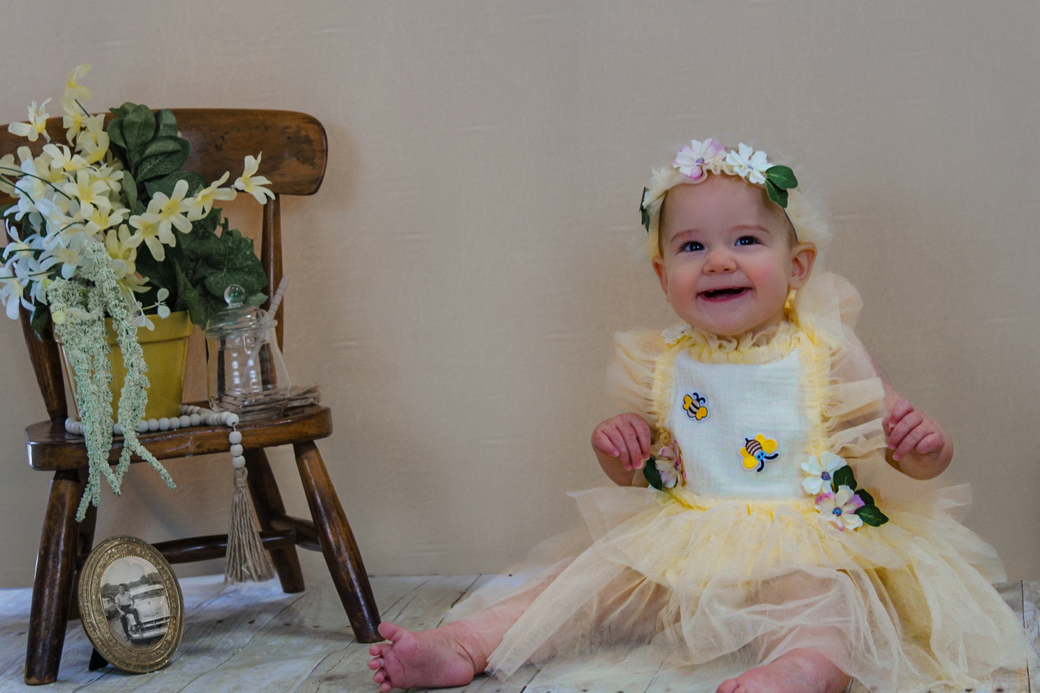 A happy baby in yellow dress sits near vintage chair with floral decorations.