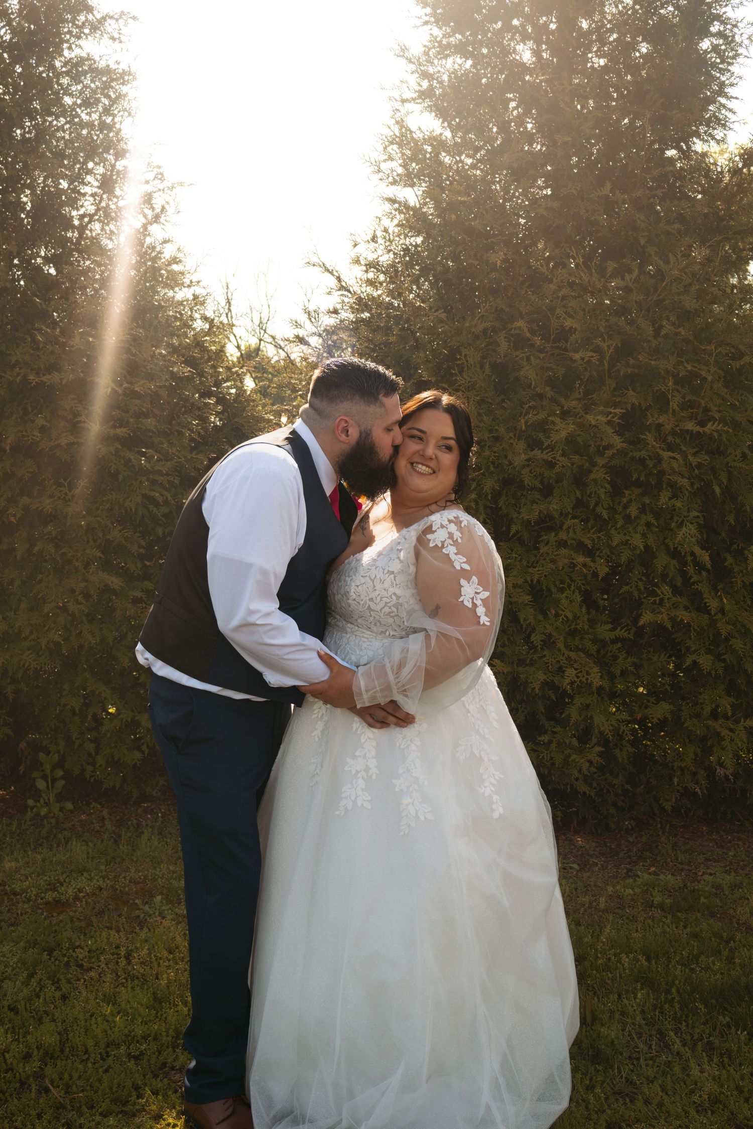 Bride in lace gown smiles as groom in vest kisses her cheek during golden hour outdoor wedding portrait.