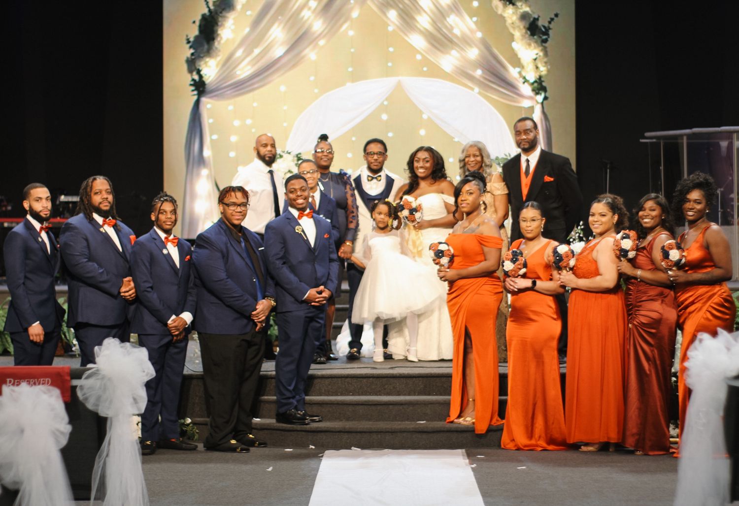 Wedding party in orange and navy attire posed under illuminated arch during ceremony.