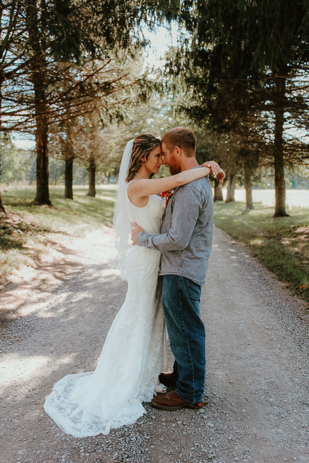 Romantic wedding couple embraces on a tree-lined gravel path in natural outdoor setting.