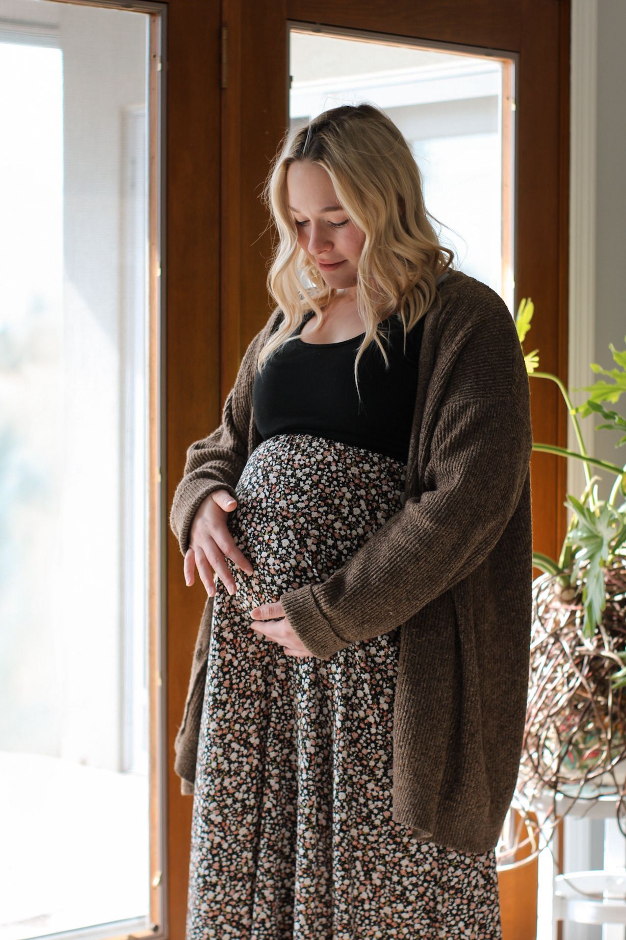 Person in floral dress and brown cardigan standing by sunlit window.