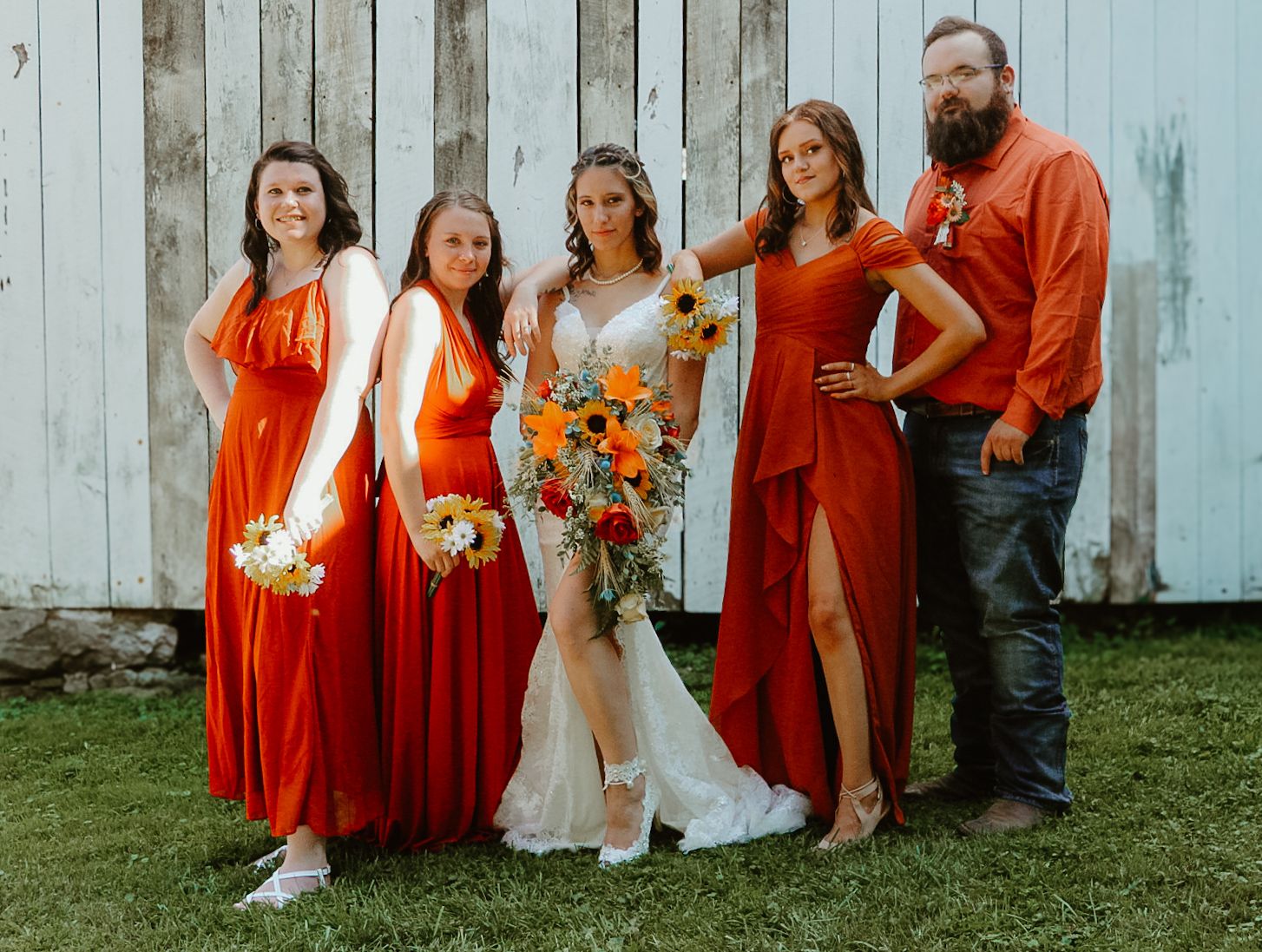 Wedding party in bright coral dresses and suits pose casually against rustic blue wooden wall with orange floral bouquets.