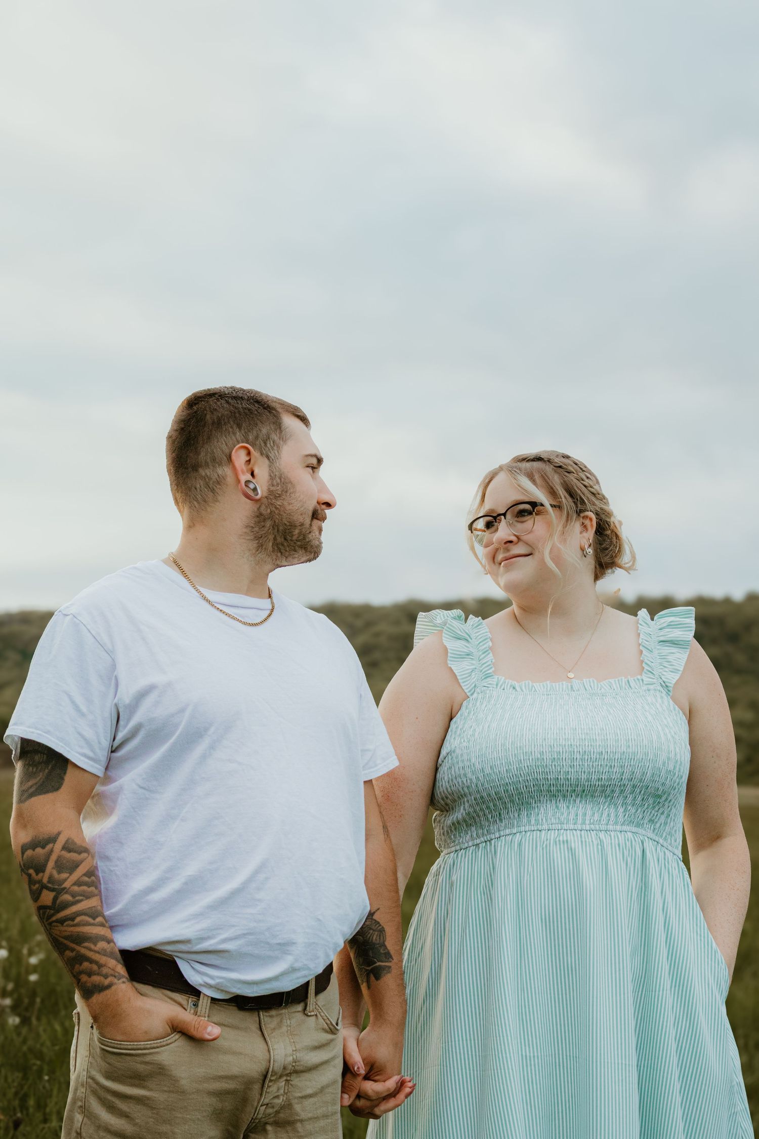 A tattooed man and a woman in a mint dress hold hands and share a loving glance in an outdoor field.