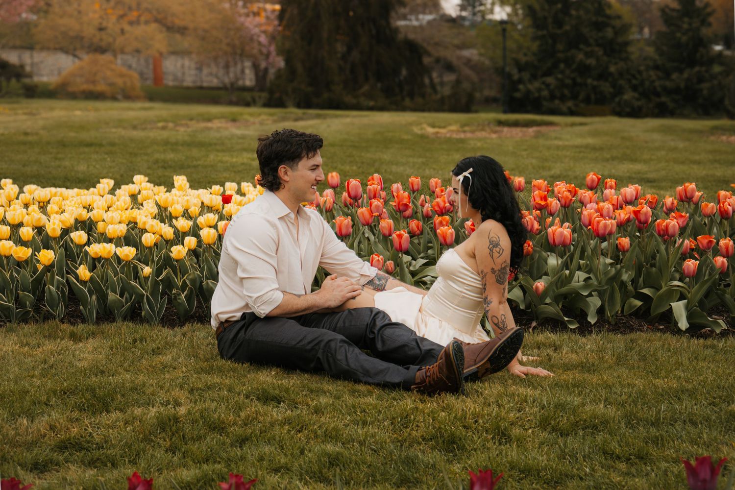 Couple sitting on grass surrounded by vibrant red and yellow tulips in a lush garden setting.