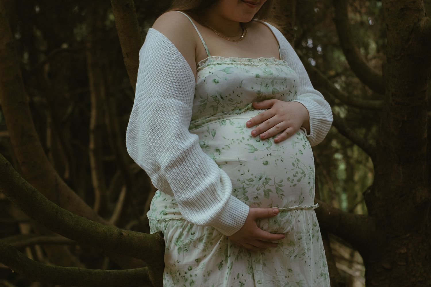 A pregnant woman in a white floral dress and sweater stands among trees in soft lighting.