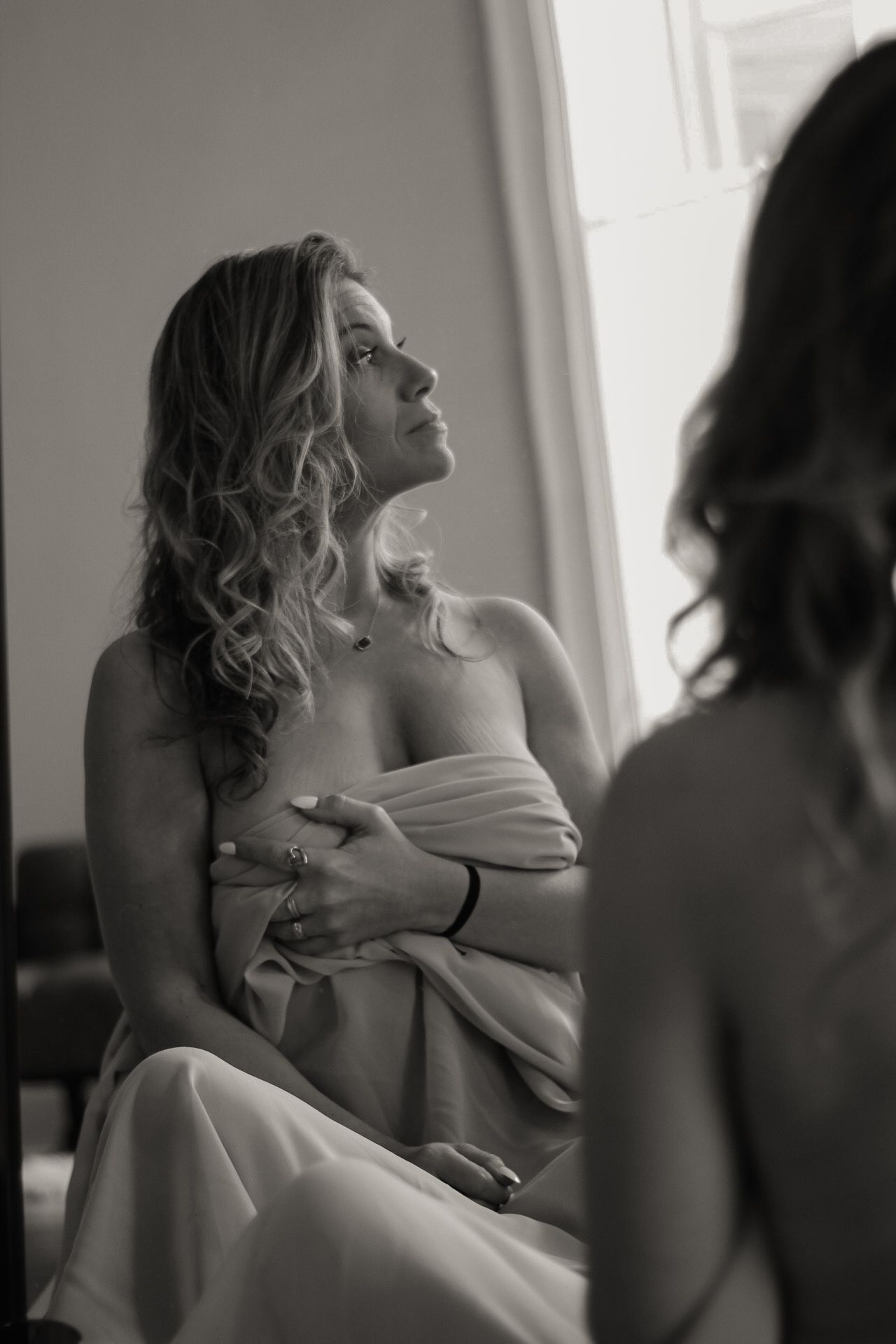 Elegant black and white artistic boudoir photo of someone in draped fabric posing thoughtfully near a mirror.