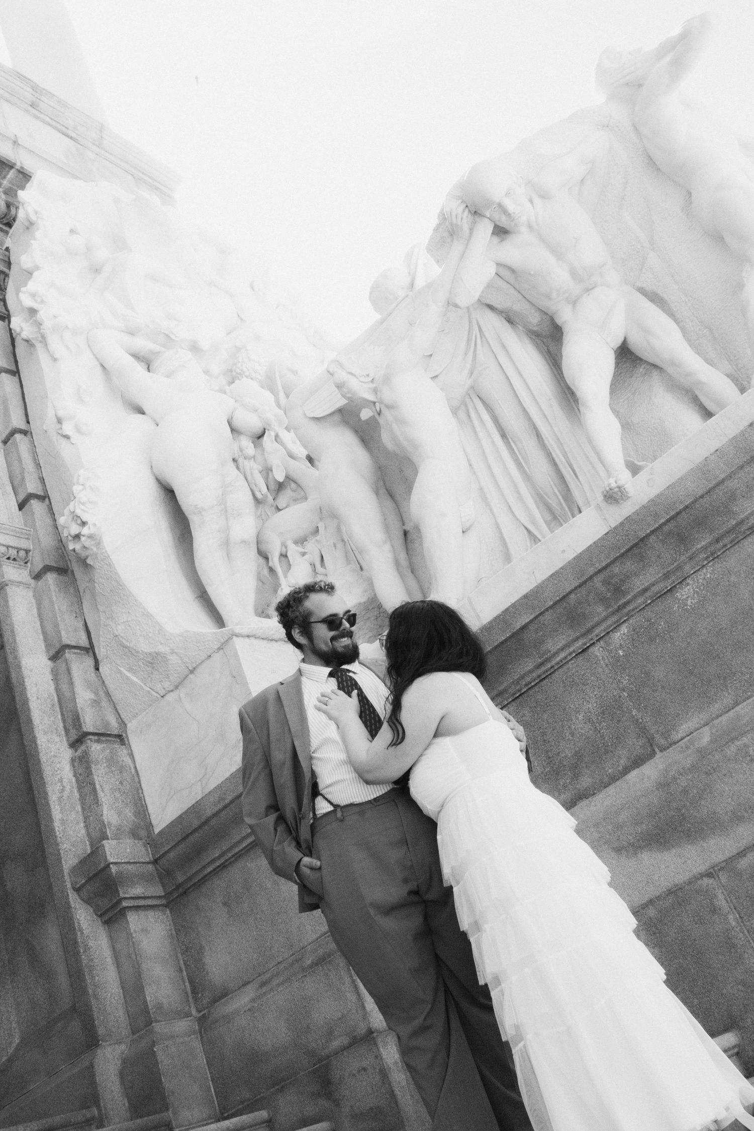 Black and white photo of bride and groom posing romantically before classical stone sculptures on a building.