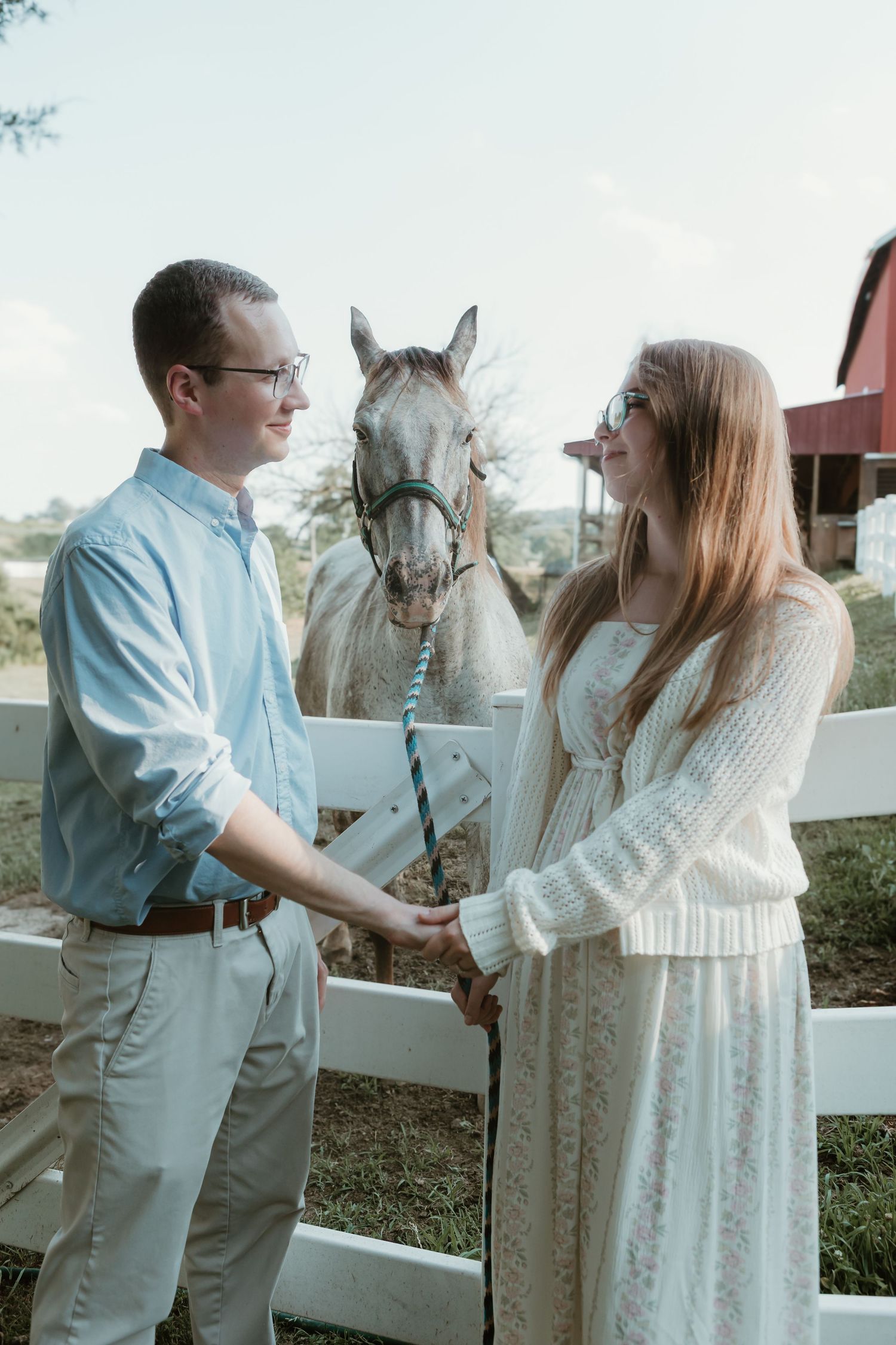 Couple holding hands and smiling at each other by a white fence with a white horse on a farm.