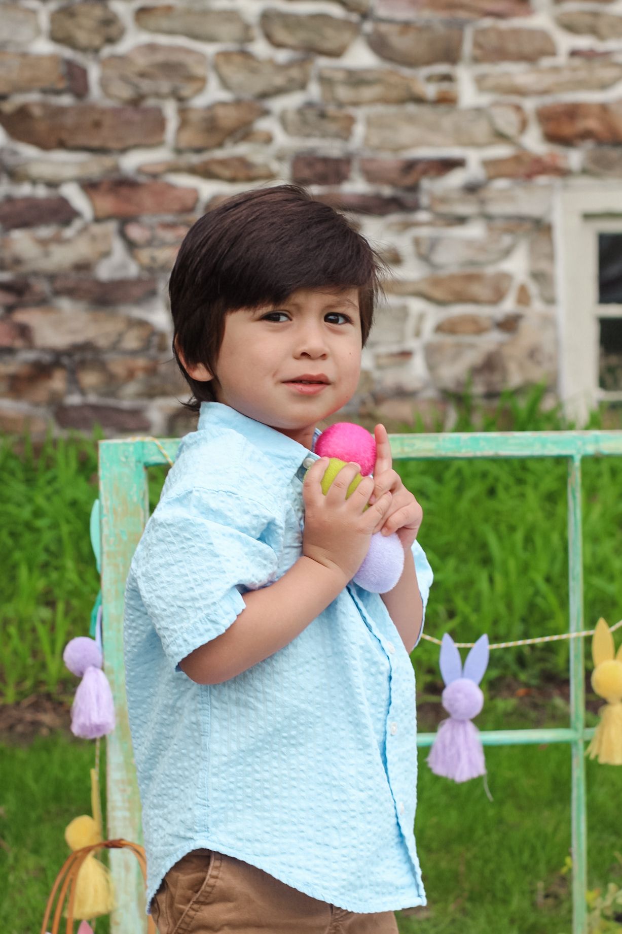 A child in a light blue shirt holds a pink Easter egg while standing by a stone wall and decorated spring backdrop.