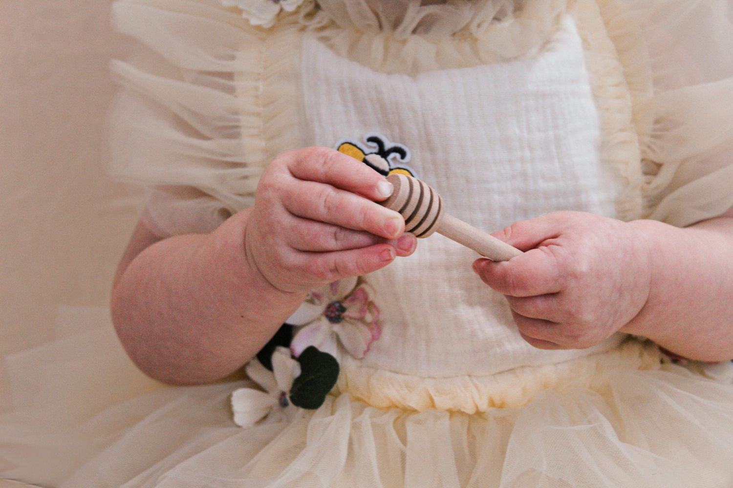 Small hands hold a toy while wearing a cream-colored ruffled dress.
