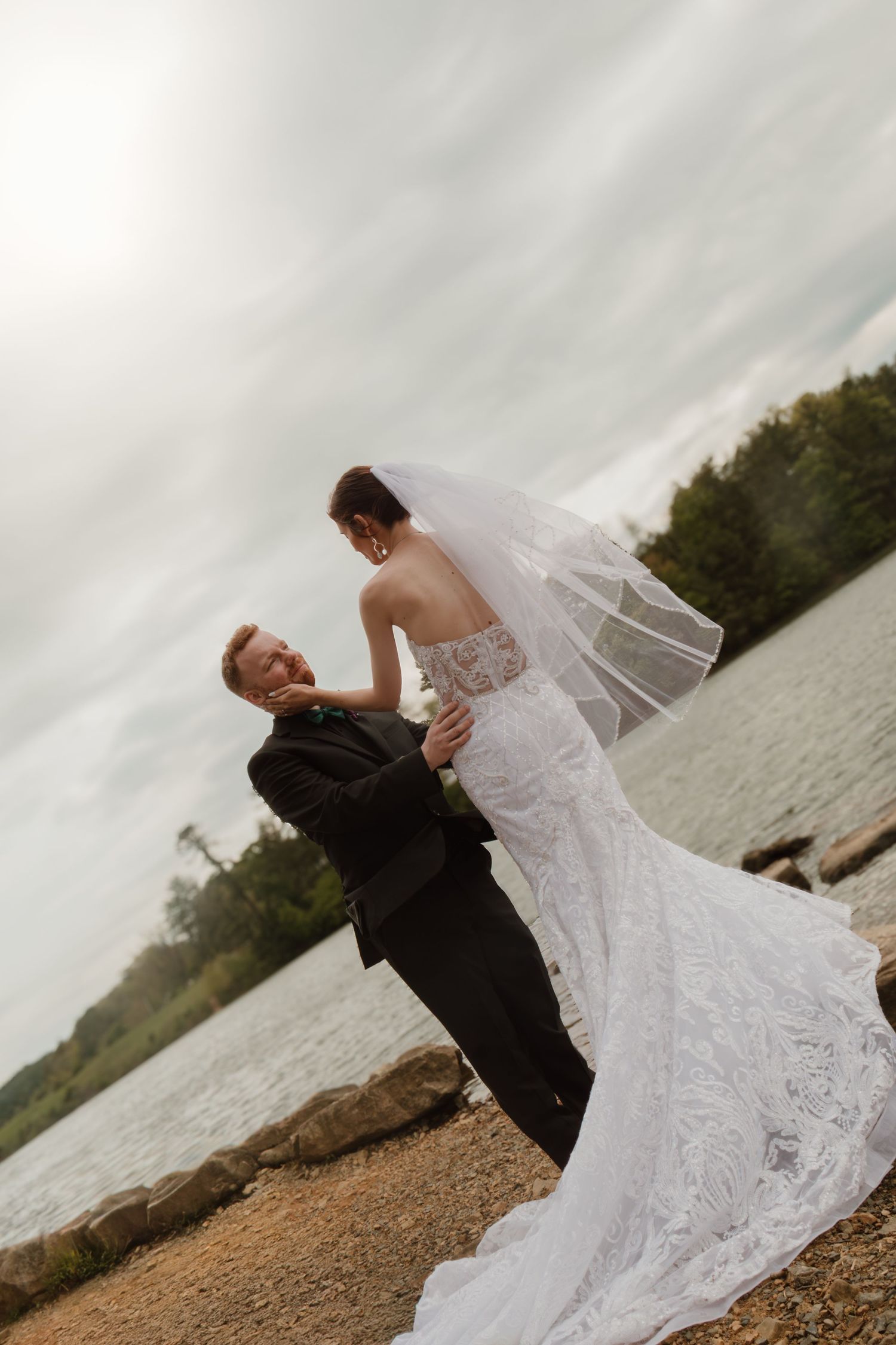 Bride in lace gown and veil dances with groom in black tux by a rocky lakeside under cloudy skies.