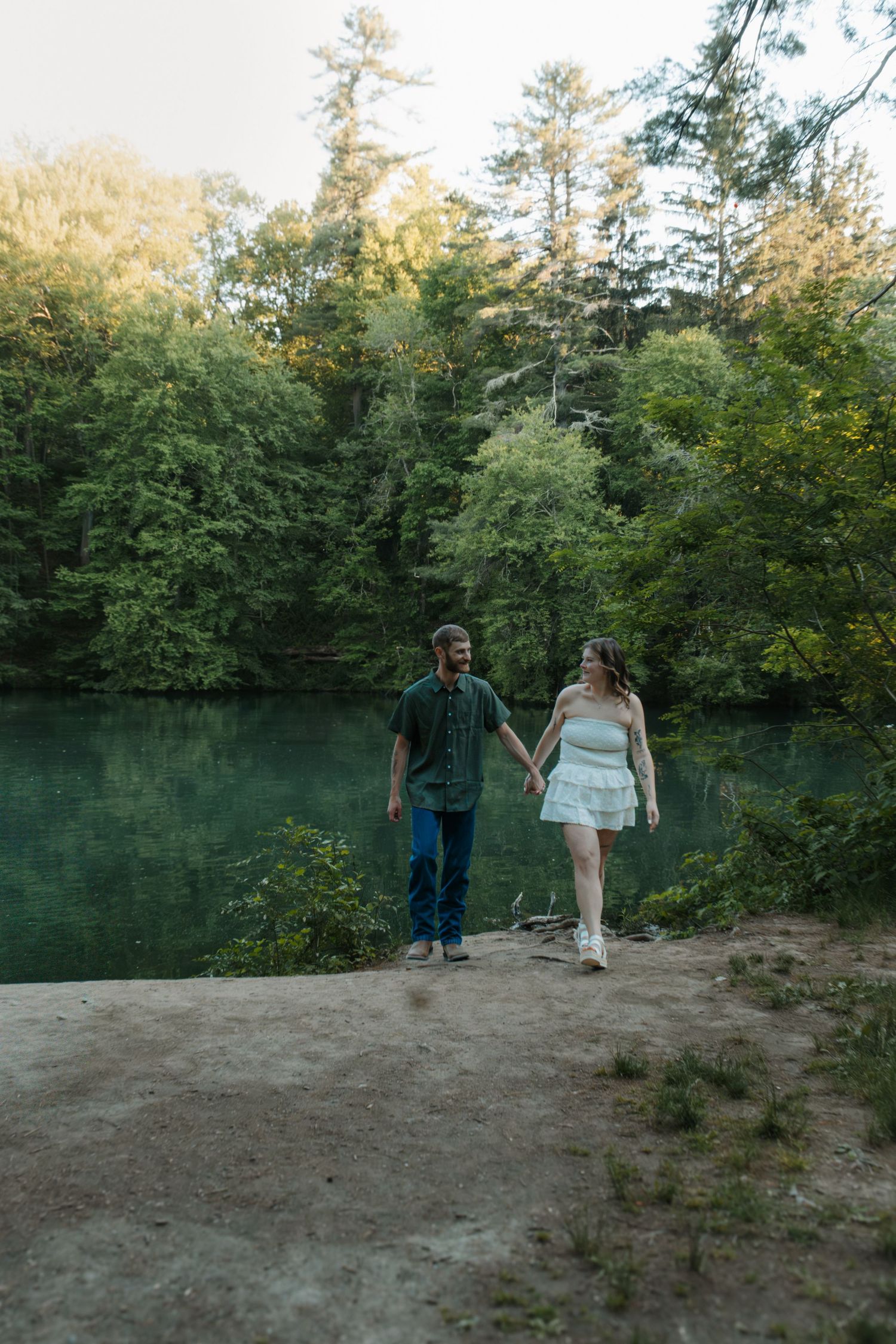 Couple holding hands walking near a serene green lake surrounded by lush forest trees during golden hour.