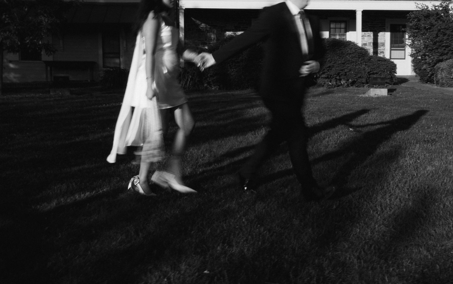 Black and white photo of a bride and groom holding hands walking on grass outside a building.