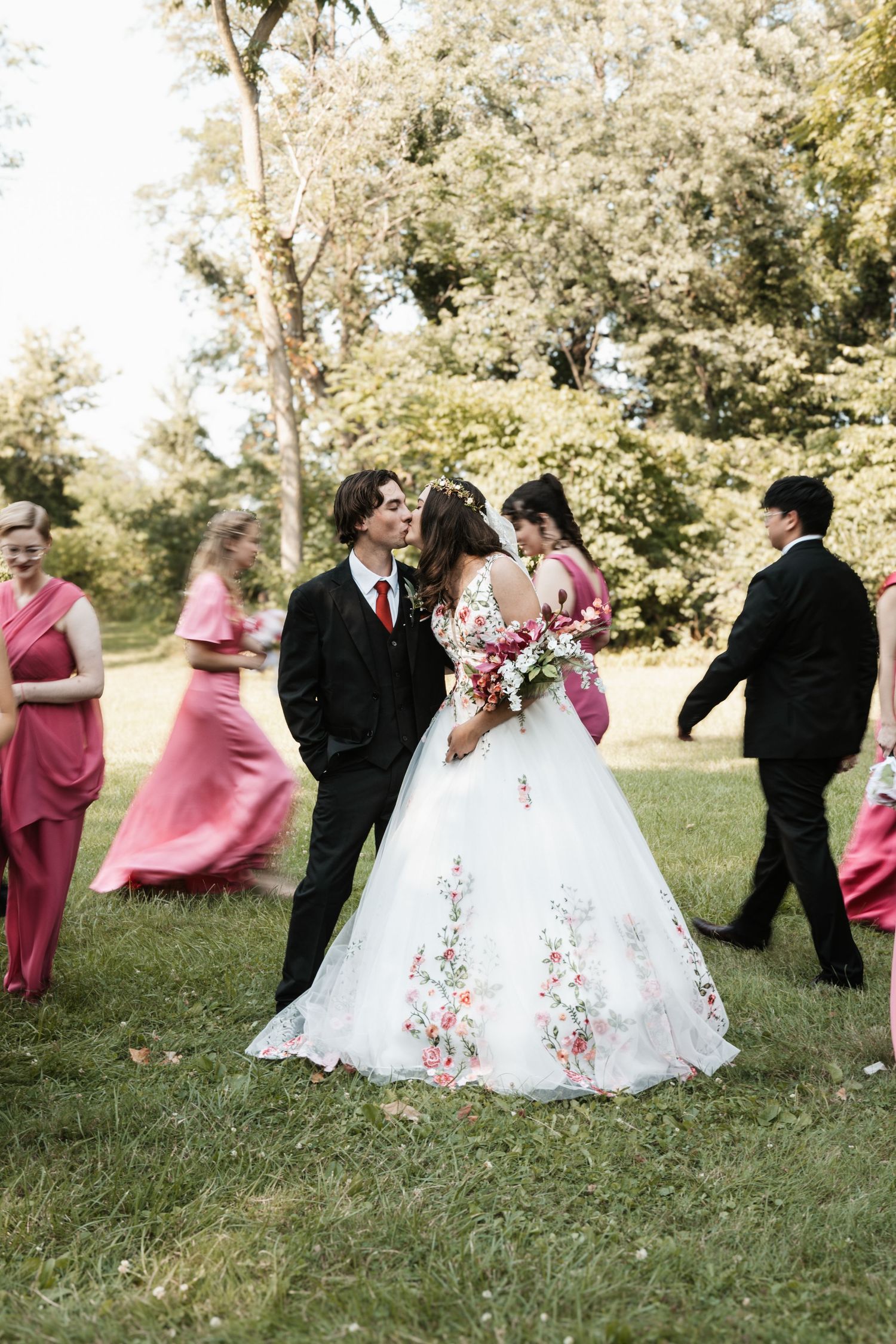 Bride and groom share a kiss in floral wedding gown outdoors, surrounded by bridesmaids in pink dresses and groomsmen.