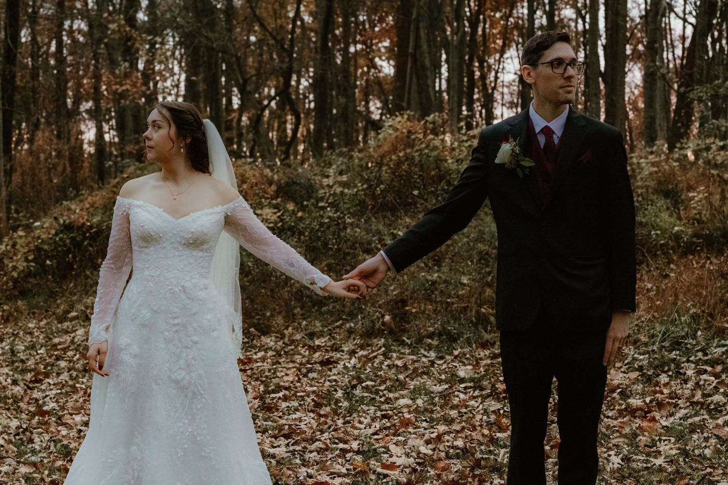 A couple in wedding attire holds hands while walking through an autumn forest with fallen leaves and bare trees.