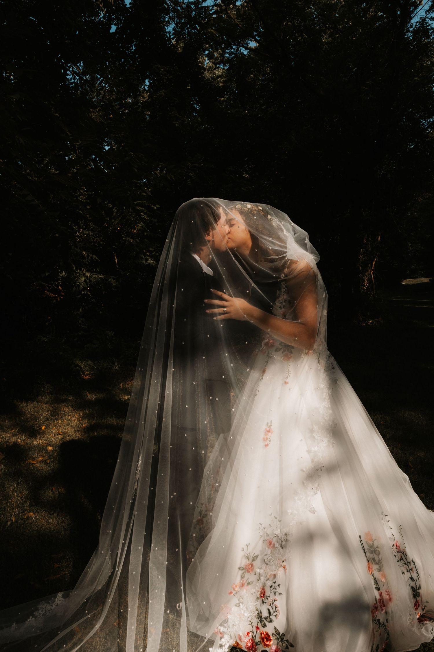 Bride and groom sharing a romantic kiss under a flowing pearl-adorned veil, wearing a floral wedding gown outdoors.