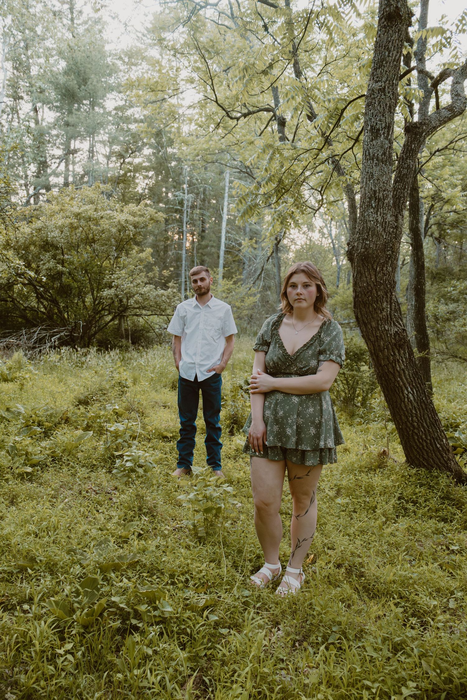 A man and woman stand in a lush green forest clearing, she in a floral romper, he in a white shirt and jeans.